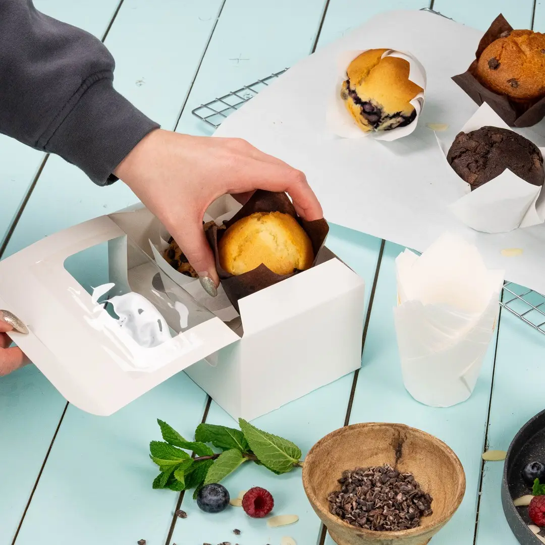 Mano che mette un muffin in una scatola bianca per cupcake con diversi prodotti da forno e ingredienti su una superficie blu.