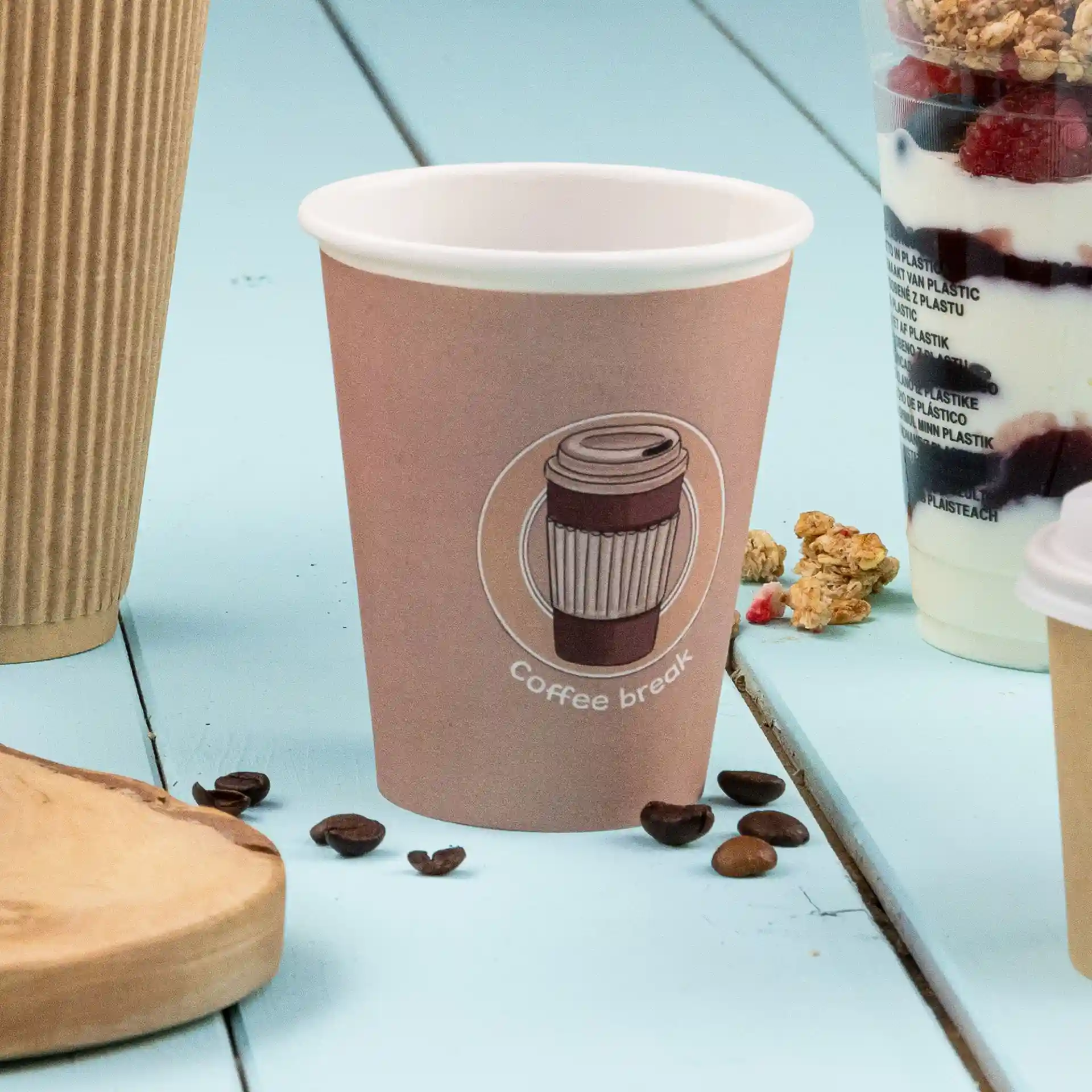 Bicchiere di carta marrone con un design di caffè stampato e testo, posato su una superficie blu con chicchi di caffè vicini.