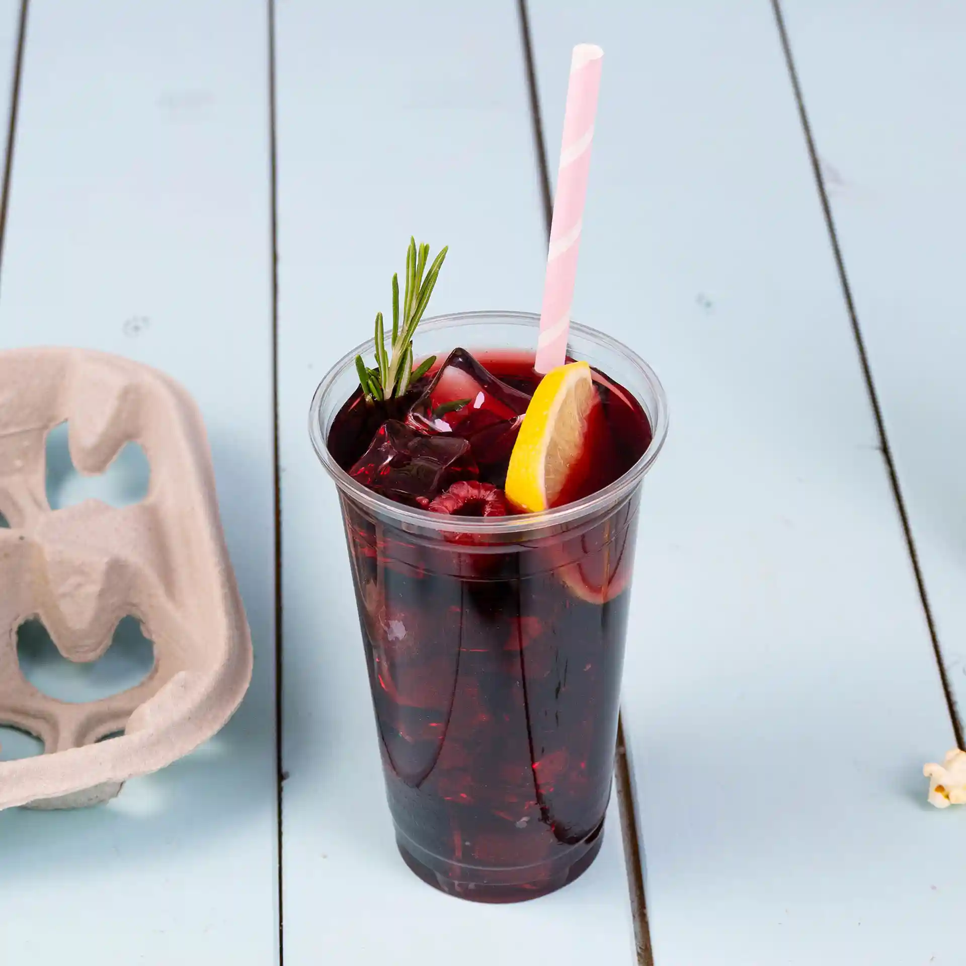 Bicchiere di plastica trasparente riempito con bevanda rossa scura, ghiaccio, fetta di limone e una cannuccia a righe rosa, con un cartone delle uova vicino.