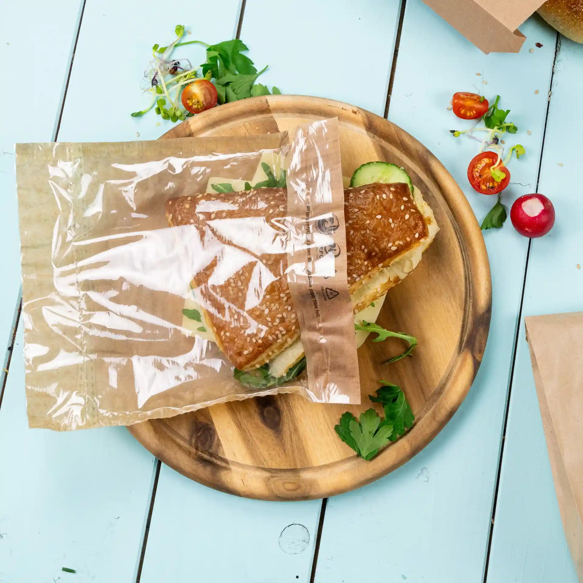 Sacchetto trasparente per snack con un panino al sesamo su un piatto di legno, circondato da erbe fresche e verdure.