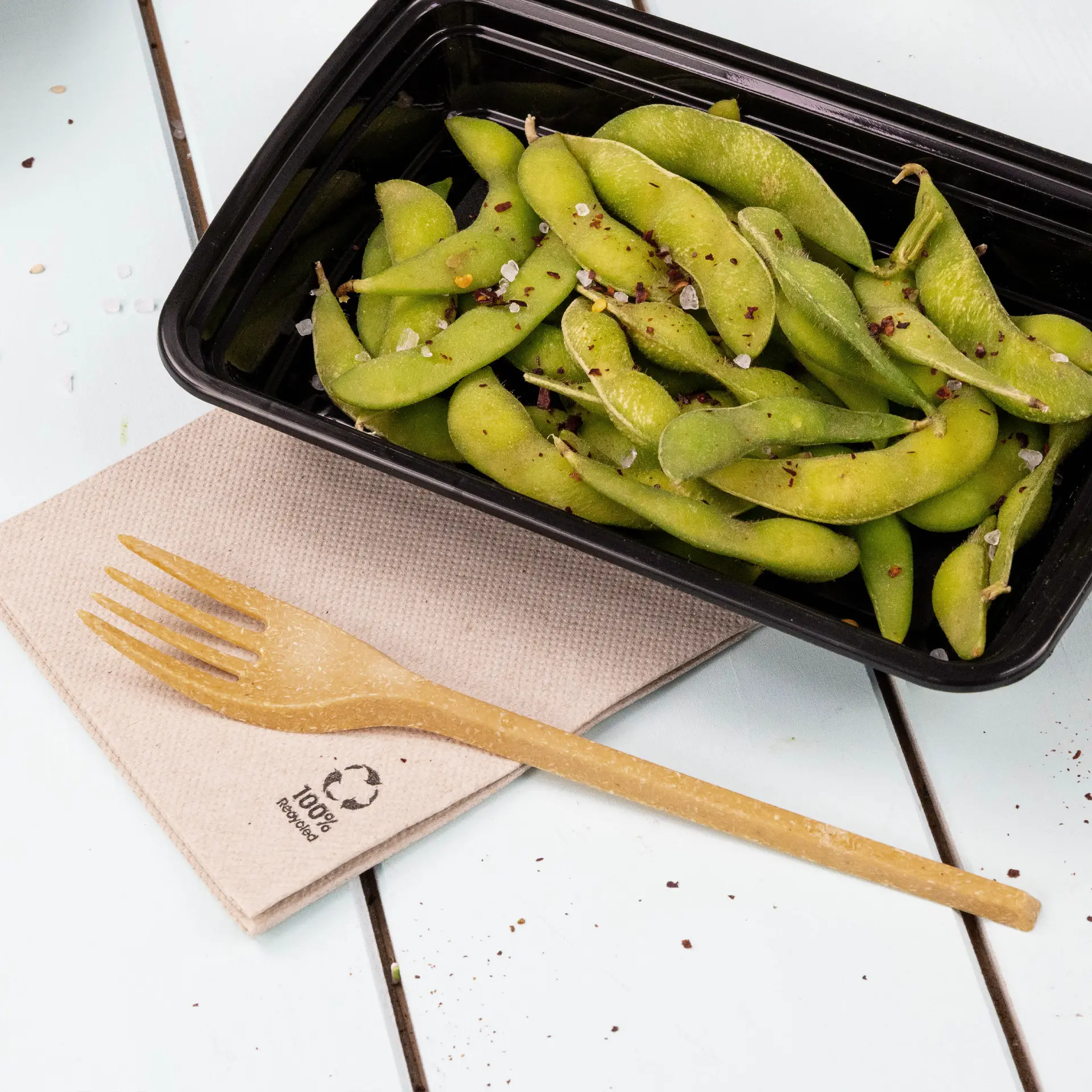 Un contenitore nero riempito di edamame verdi guarnito con spezie, accompagnato da una forchetta biodegradabile su un tovagliolo di carta riciclata.