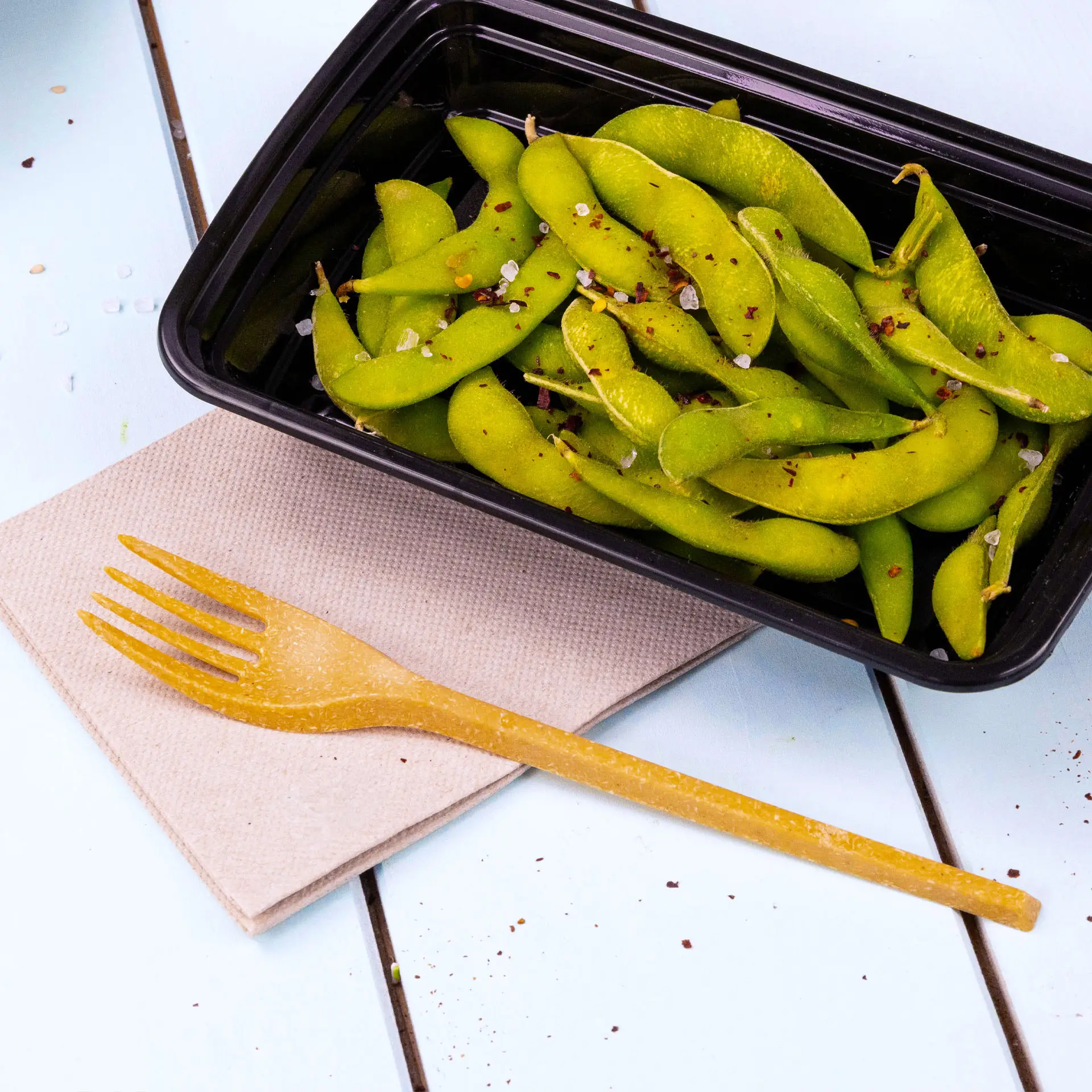 Immagine di un contenitore nero pieno di fagioli edamame verdi, una forchetta gialla su un tovagliolo beige accanto.