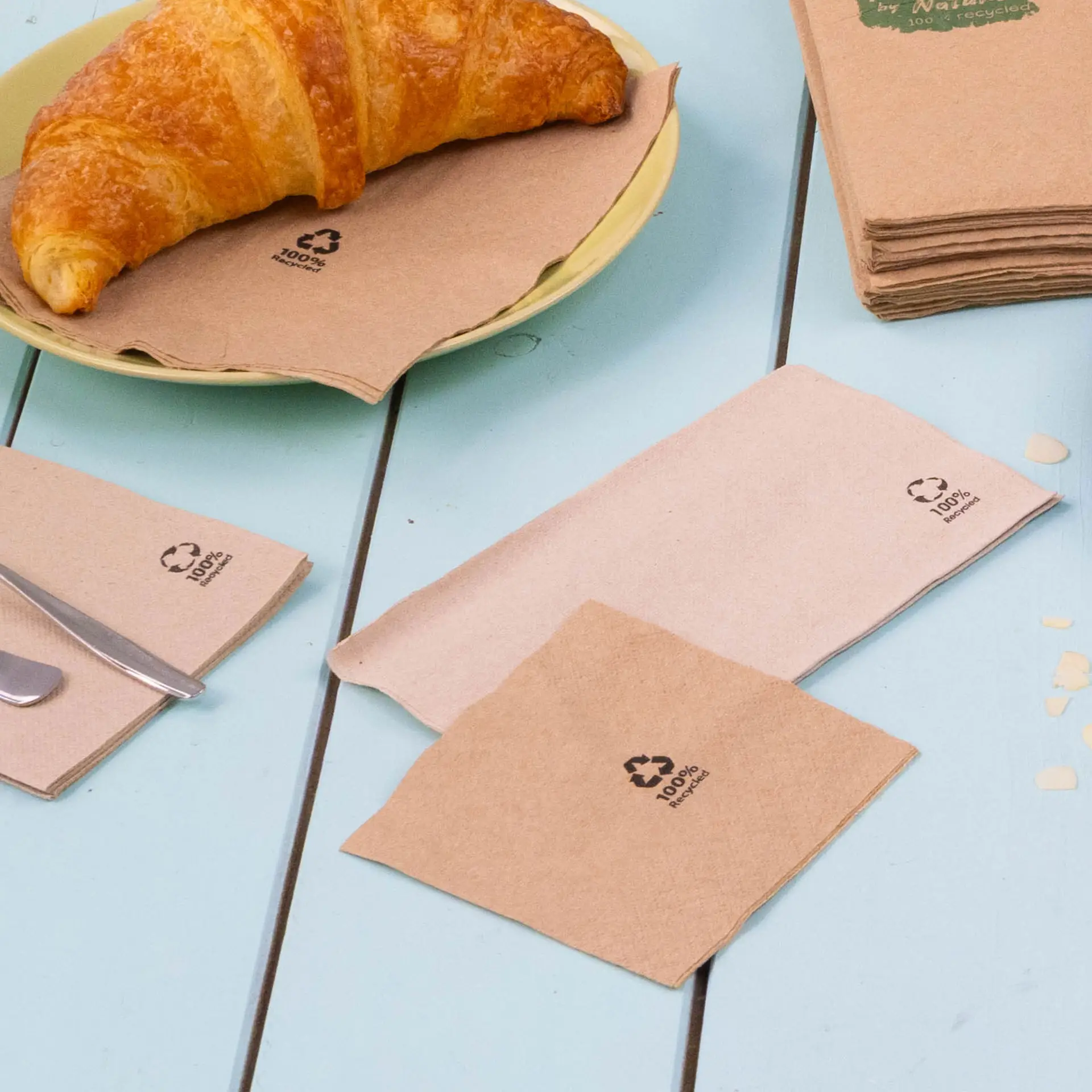 Immagine di tovaglioli per cocktail biodegradabili marroni su un tavolo blu, con un cornetto su un piatto, mostrano simboli di riciclaggio.