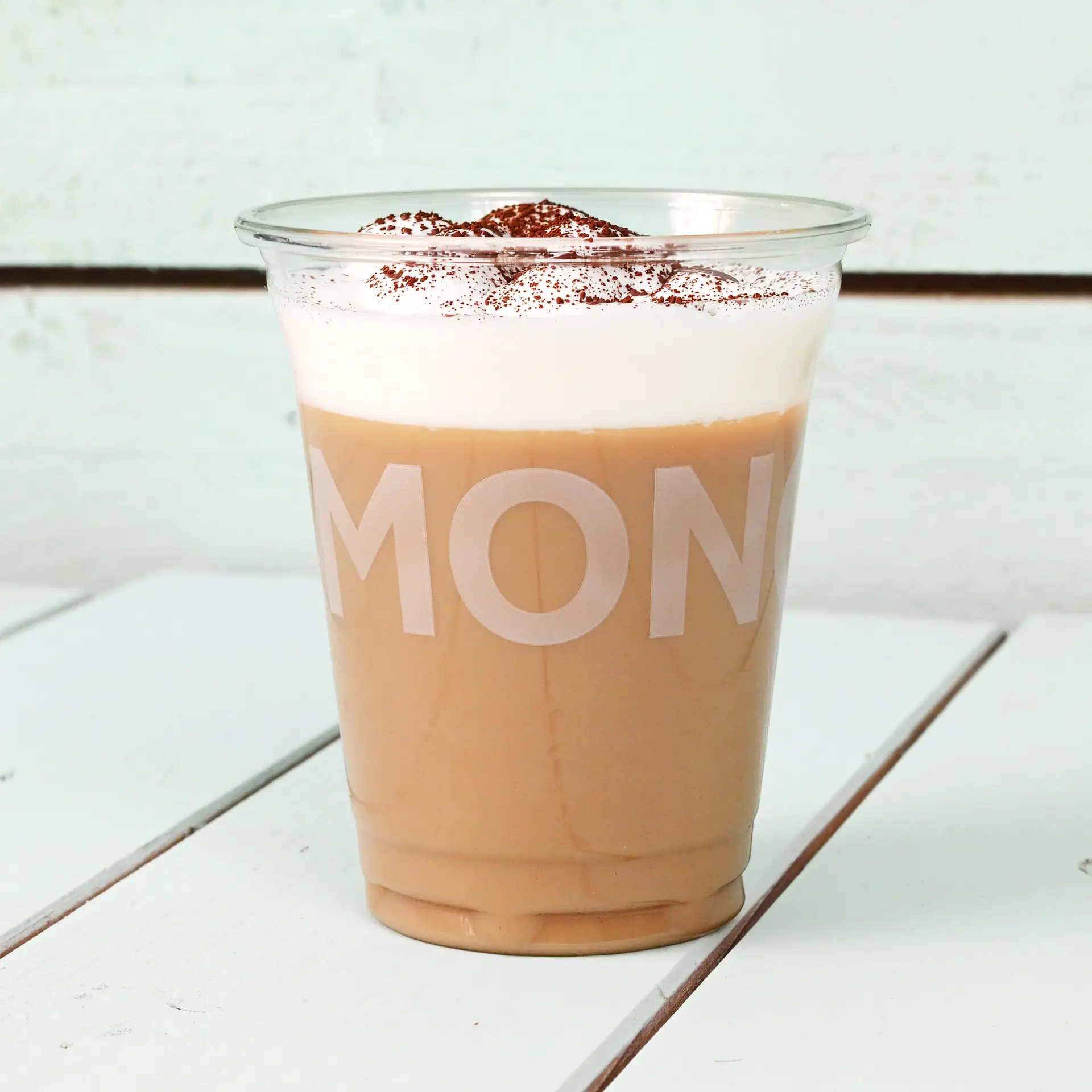 Bicchiere PLA trasparente da 12 oz con bevanda ghiacciata guarnita con schiuma e cacao, su una superficie in legno bianco.