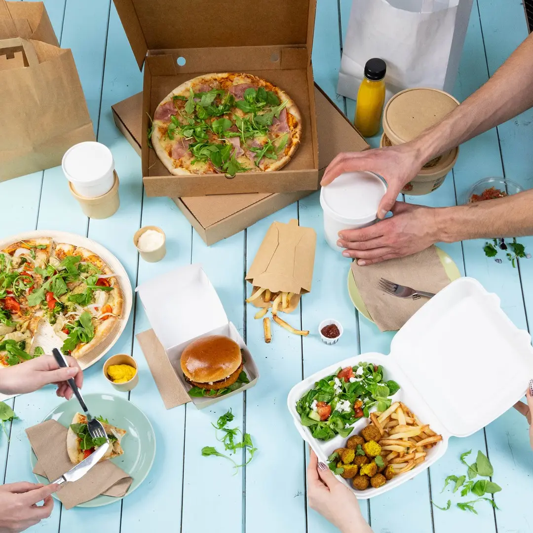Tavolo apparecchiato con vari cibi da asporto, tra cui pizze, un hamburger, patatine, insalata e bevande in contenitori.