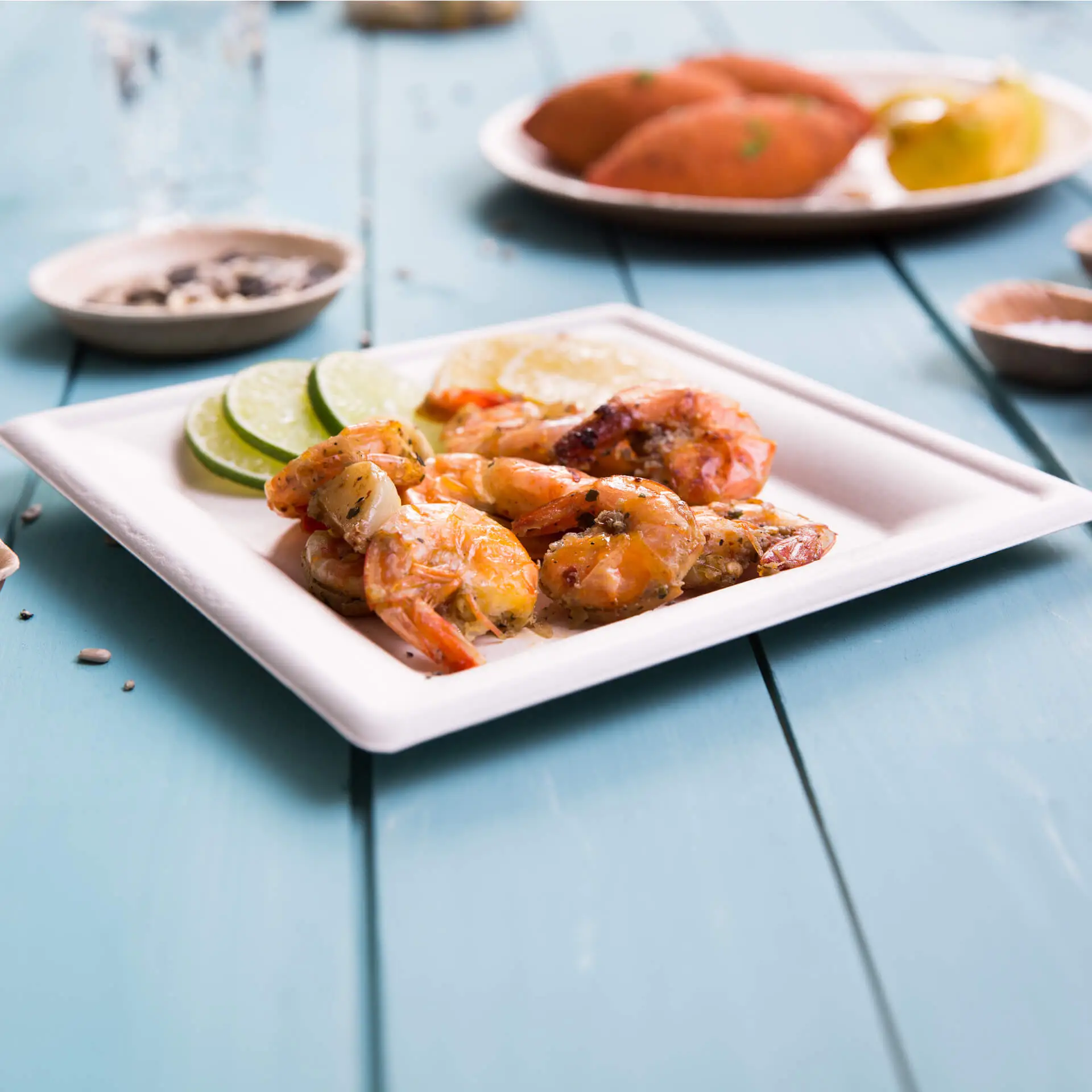 Gamberetti grigliati con fette di lime su un piatto biodegradabile di canna da zucchero, serviti su un tavolo di legno blu.