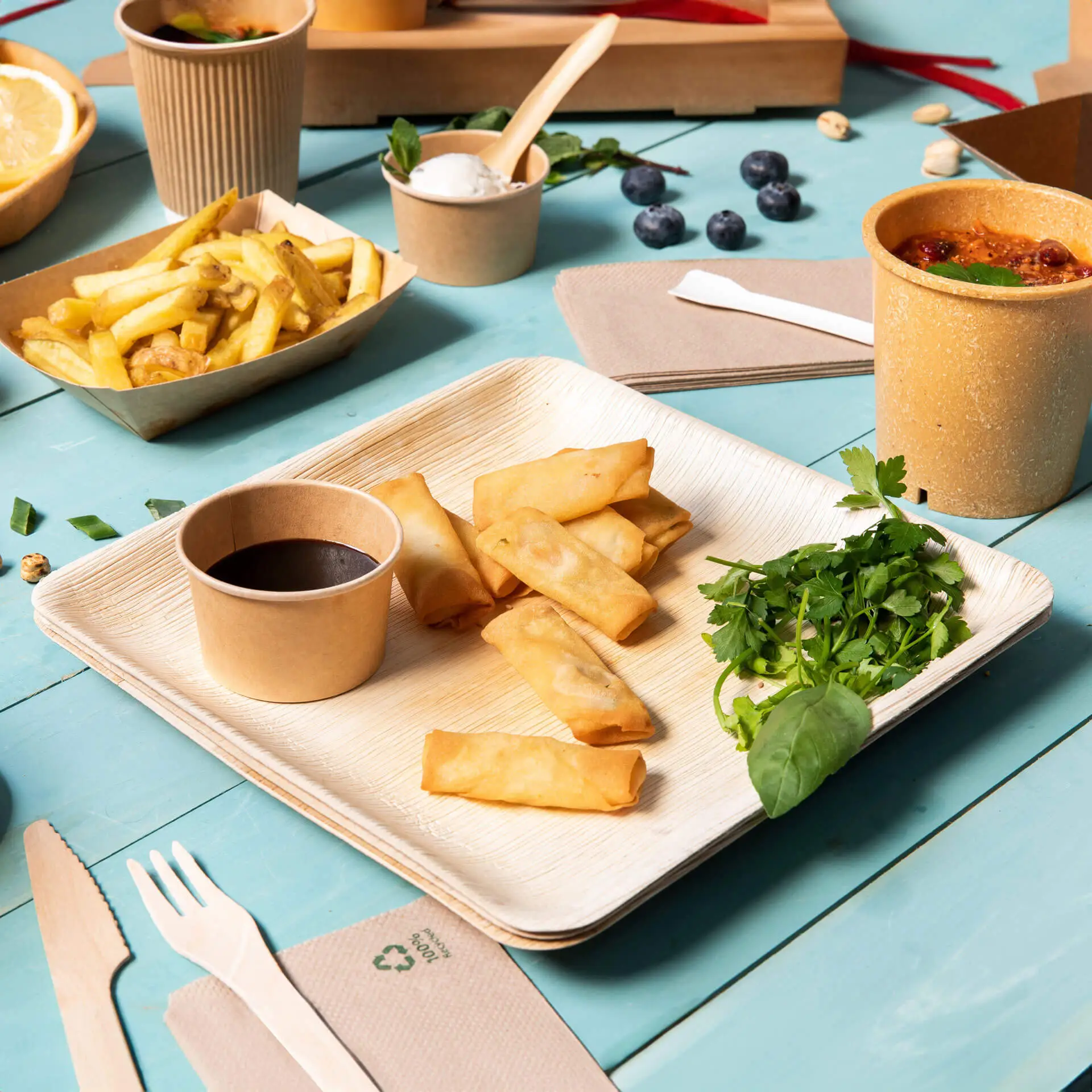 Un piatto quadrato in foglia di palma con involtini primavera, una ciotola di salsa e erbe fresche; servito su un tavolo blu con contorni.