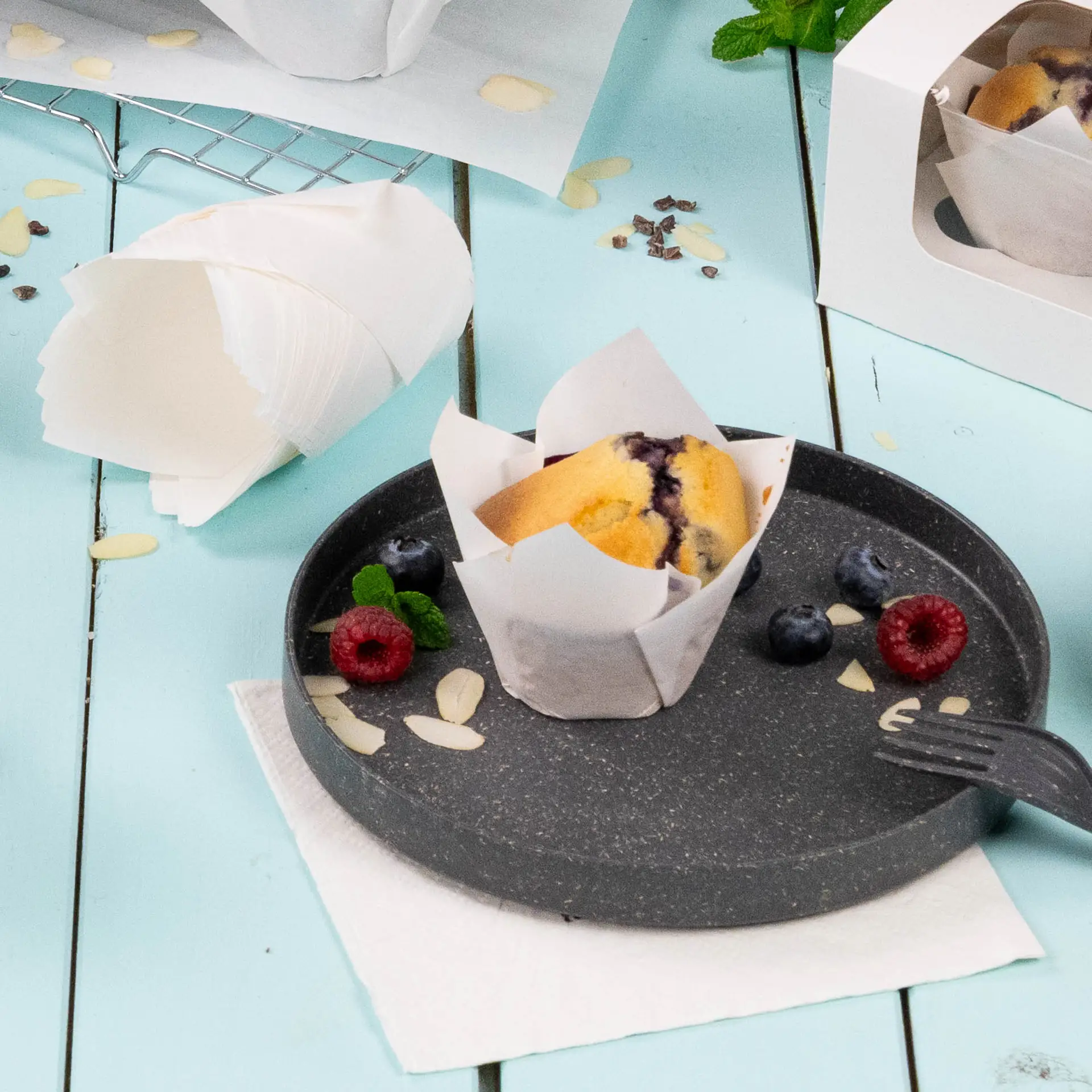 Un pirottino bianco con carta anti-grasso su un piatto scuro; guarnito con lamponi, mirtilli e fette di mandorla.