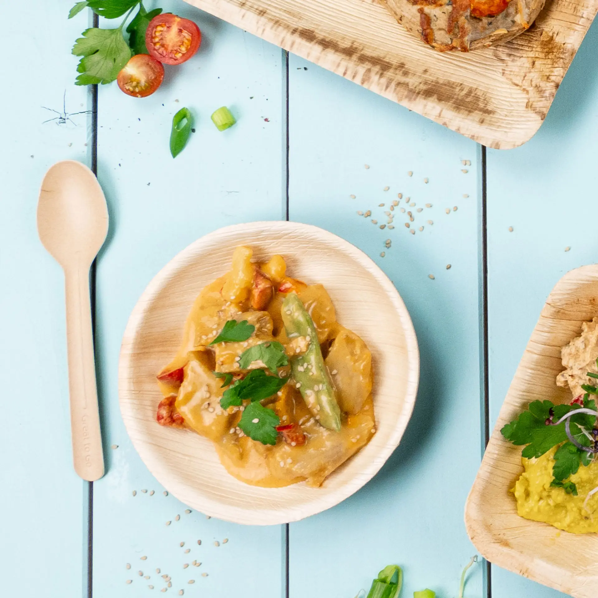 Piatto rotondo con piatto cremoso a base di verdure e sesamo; cucchiaio di legno accanto; sfondo azzurro con pomodorini e cipollotti.