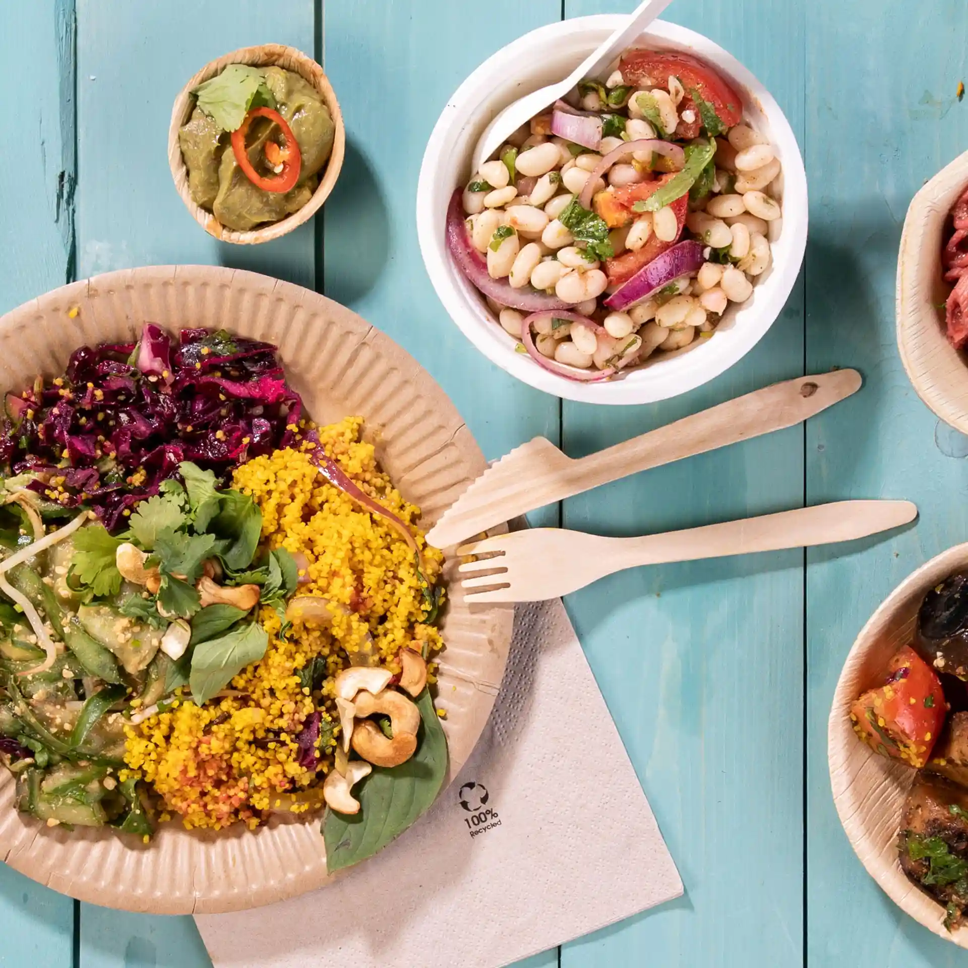 Immagine di un piatto con cibo colorato, tra cui couscous, insalata e guarnizioni, accompagnato da forchette di legno e piccole ciotole di condimenti.