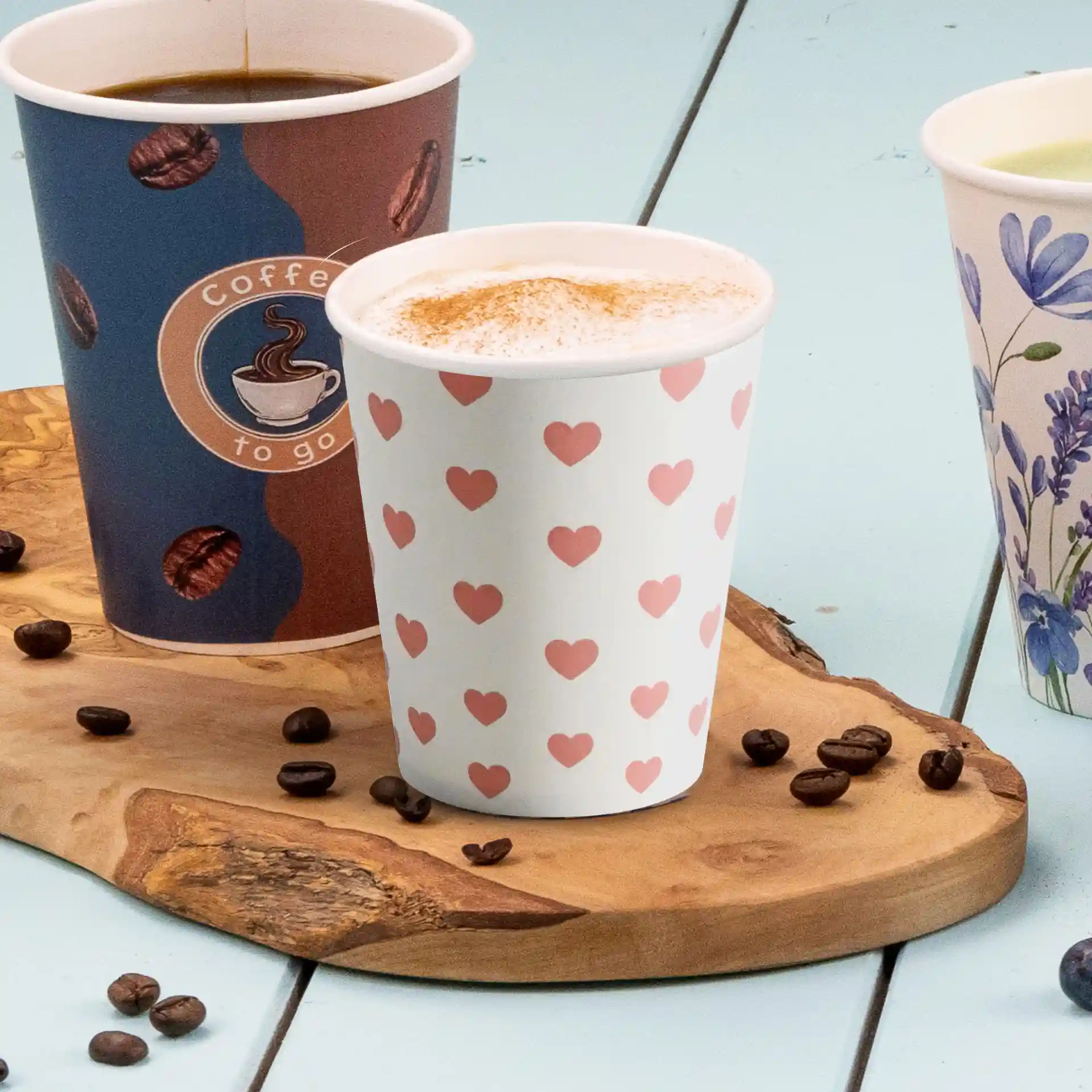 Tre bicchieri di carta su un tavolo di legno, uno con motivo a cuore, uno con logo di caffè e uno con design floreale.