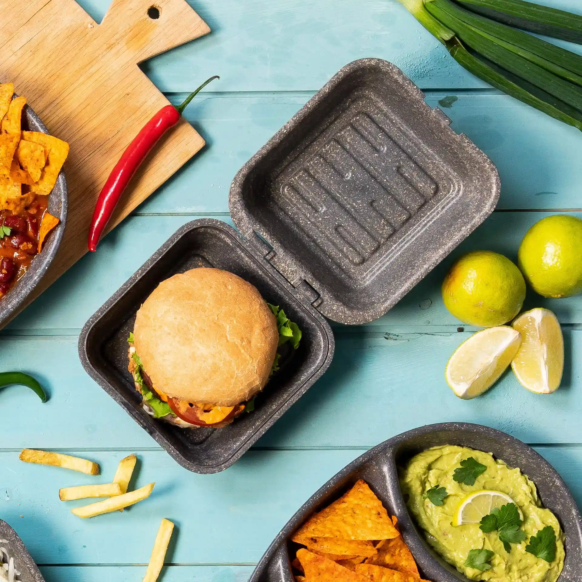 Scatola per hamburger grigia riutilizzabile con coperchio, contenente un hamburger, circondato da lime, nachos e un peperoncino.