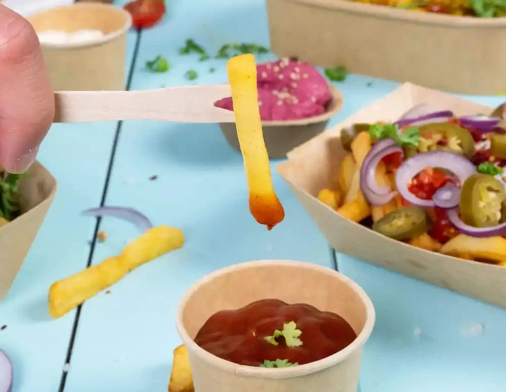 Una mano tiene un bastone di legno con una patatina sopra un bicchiere di ketchup posato su una superficie blu.