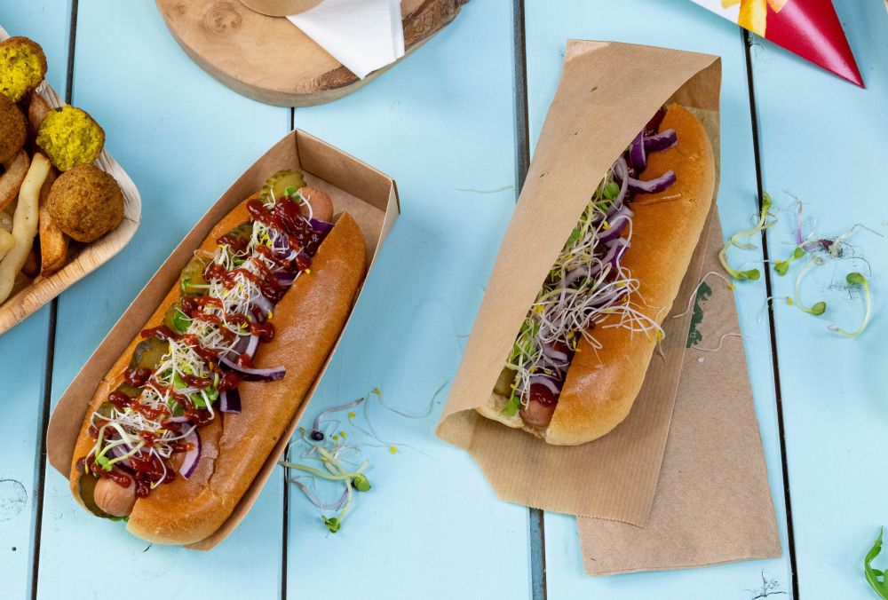 Due hot dog con vari condimenti, tra cui cetrioli, cipolle e germogli, serviti in contenitori di carta su un tavolo blu.