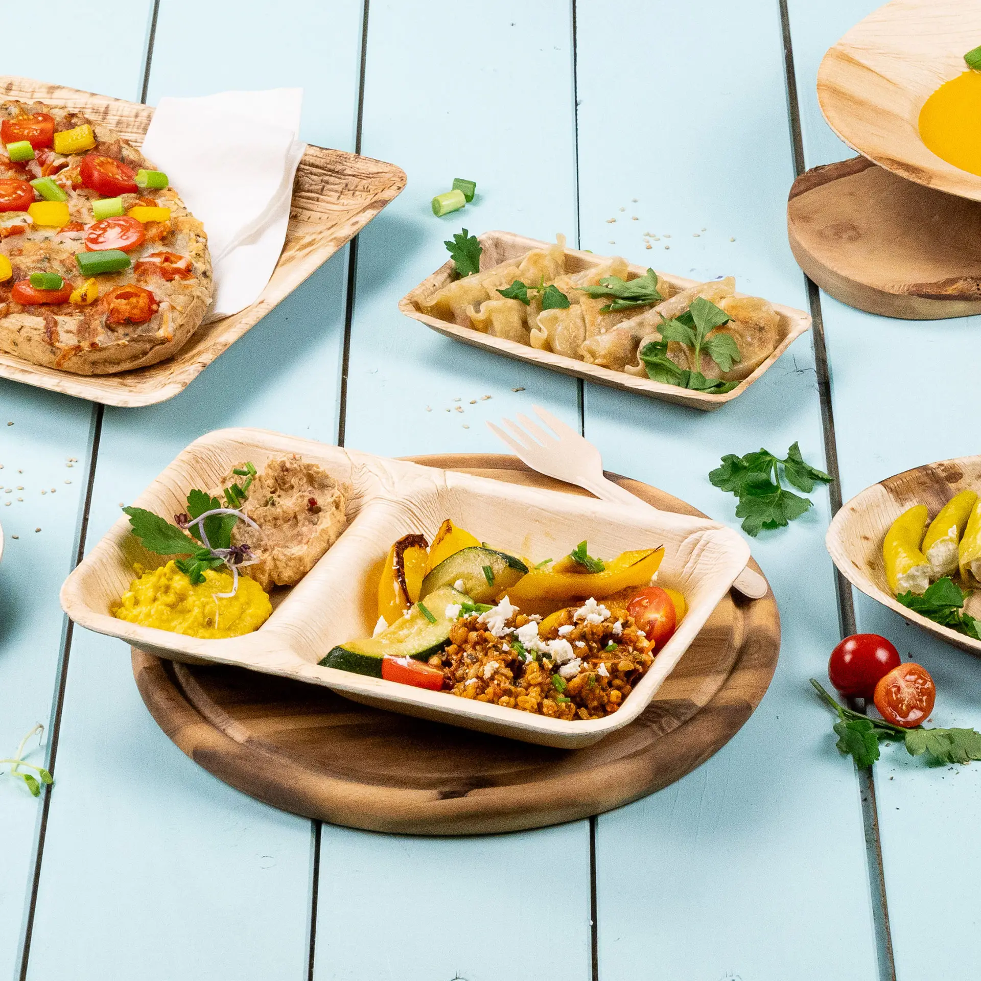 Piatto da menu a tre scomparti in foglia di palma con hummus, verdure saltate e ravioli su sfondo blu chiaro.