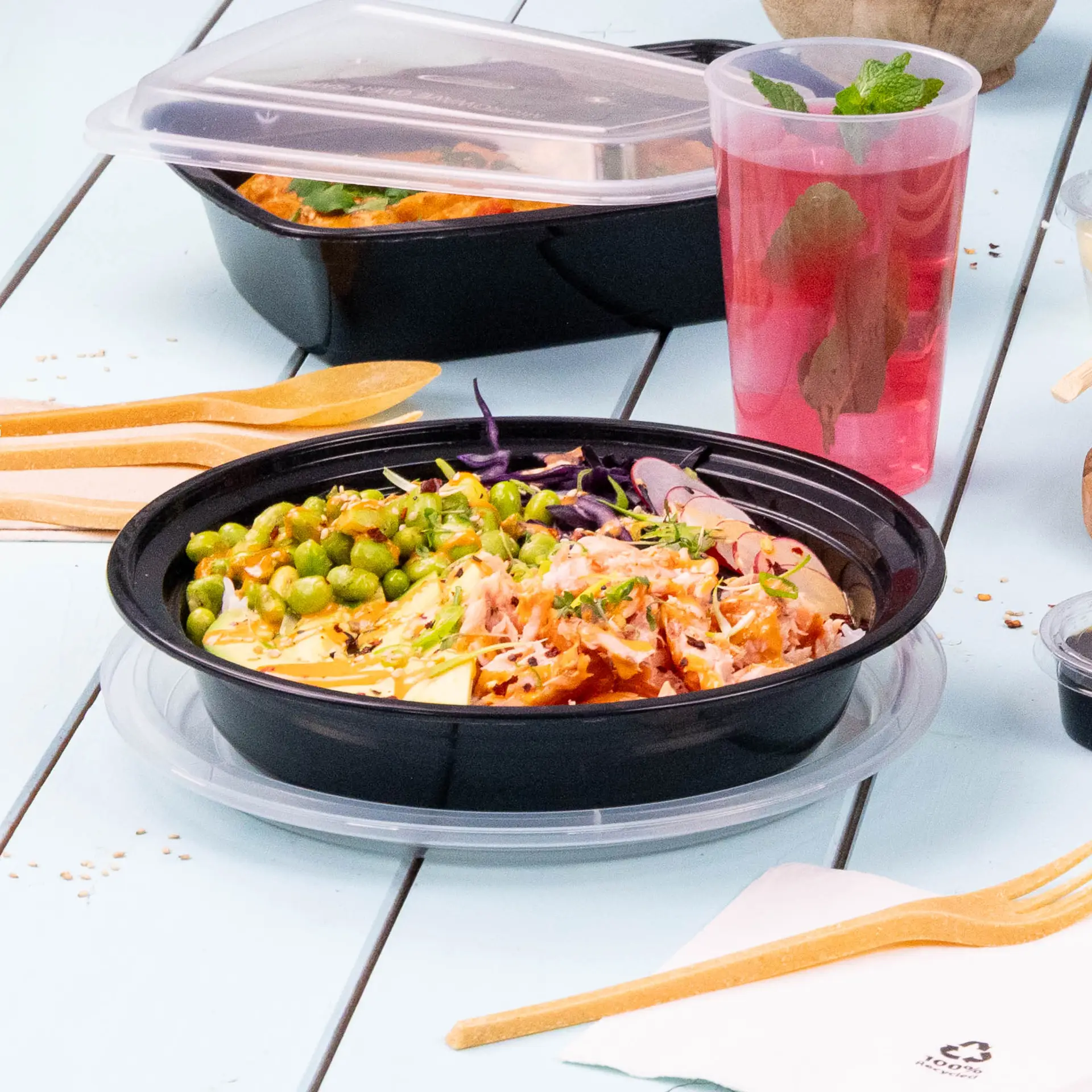 Un piatto nero riutilizzabile con cibo misto, tra cui piselli e verdure grattugiate, su un tavolo azzurro; posate in legno vicine.