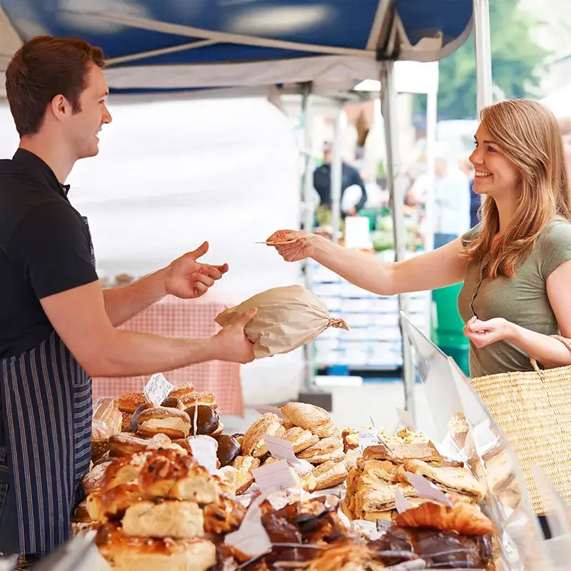 Un venditore porge una busta di carta marrone a un cliente presso un banco di mercato con vari prodotti da forno.
