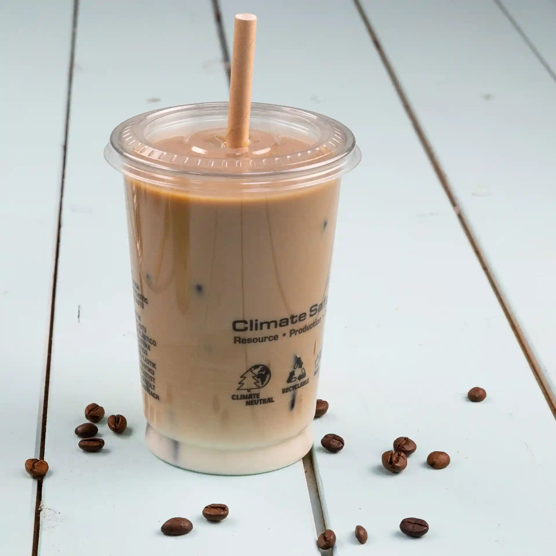 Bicchiere trasparente con caffè freddo e una cannuccia di carta, posato su una superficie azzurra con chicchi di caffè sparsi.
