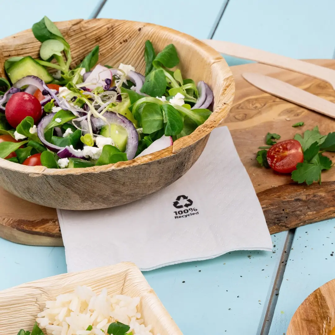 Insalata in una ciotola di legno con un tovagliolo etichettato "100% Recycled", accanto a riso e un pomodorino su un tagliere di legno.