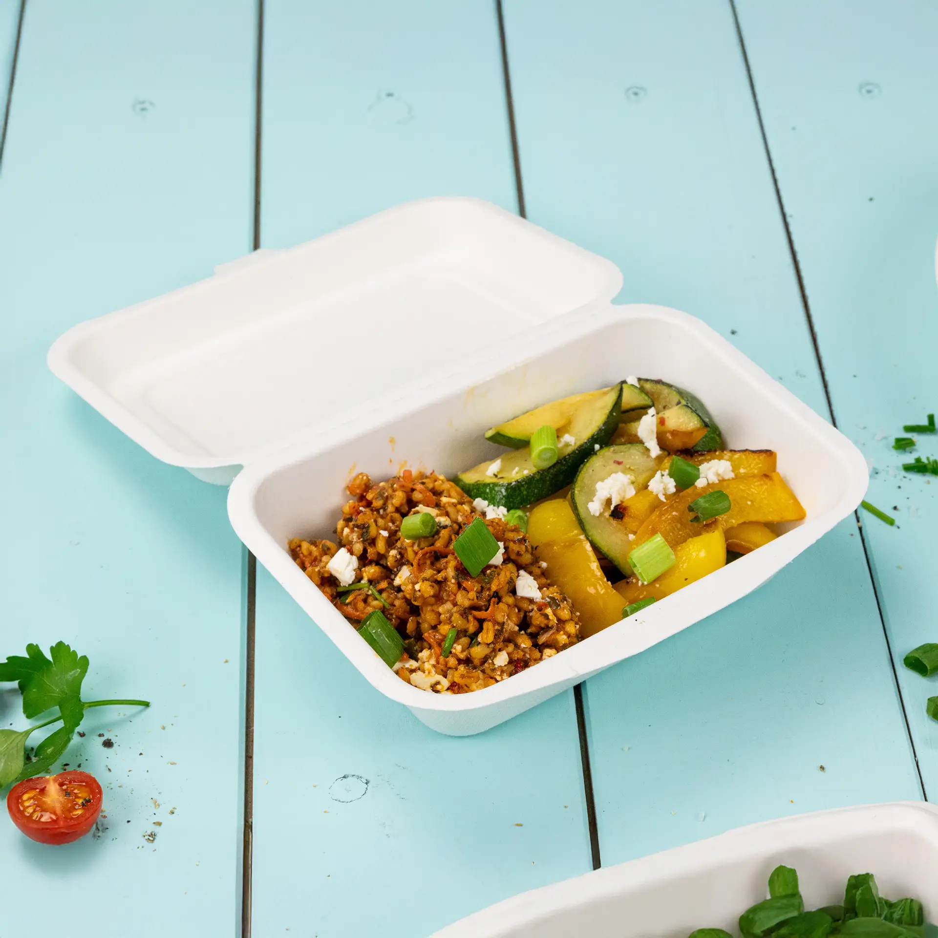 Un contenitore per alimenti biodegradabile in canna da zucchero con coperchio a cerniera, ripieno di cereali e verdure miste, su uno sfondo blu.