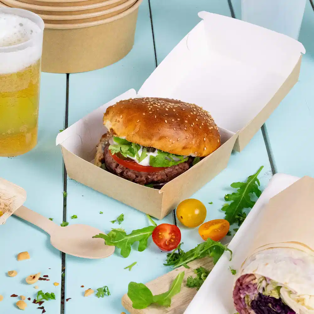 Un hamburger con panino al sesamo, lattuga, pomodoro e salsa in una scatola da asporto marrone, accompagnato da verdure e birra.