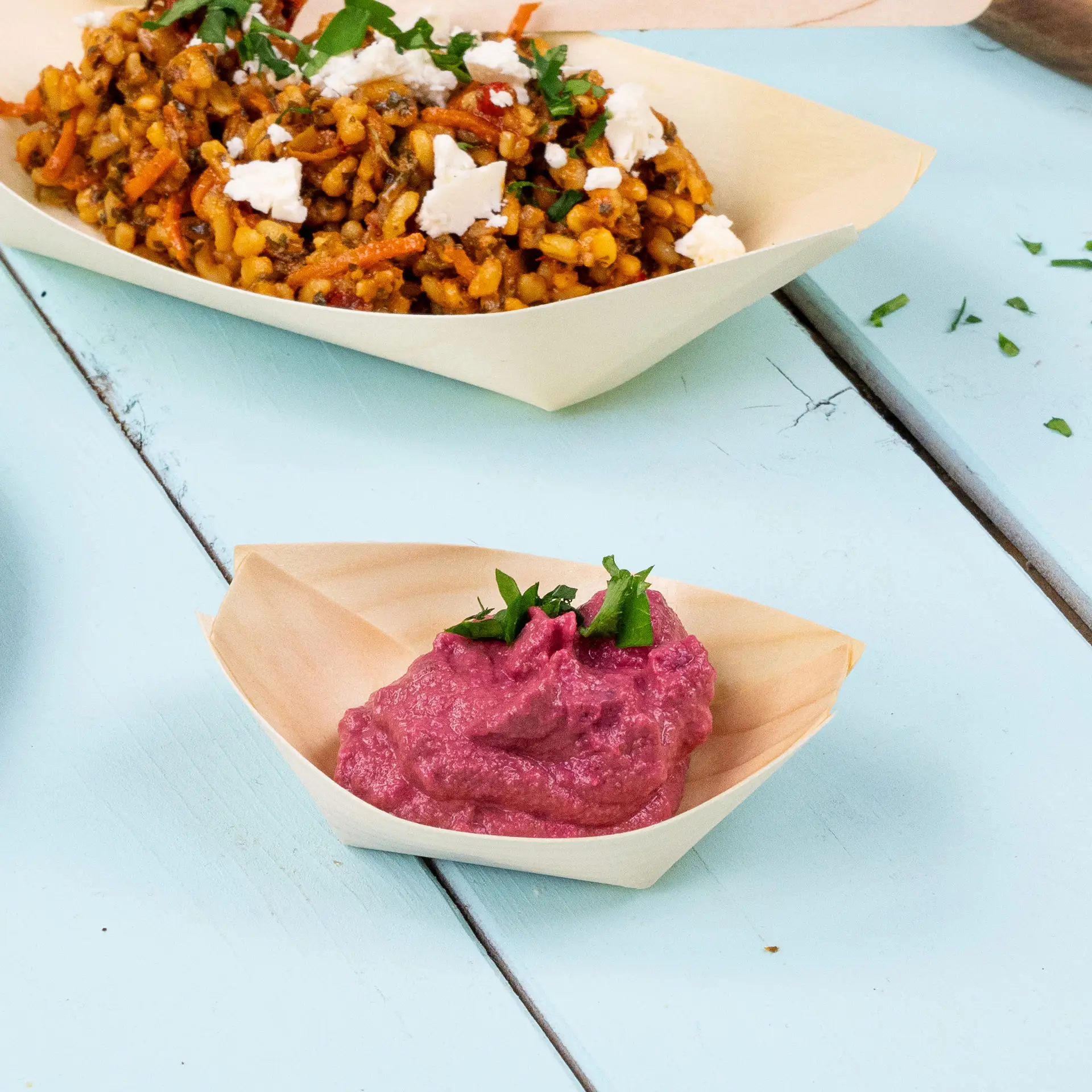 Ciotola di legno a forma di barca con salsa rosa guarnita con erbe verdi, accanto a un piatto di cereali misti.