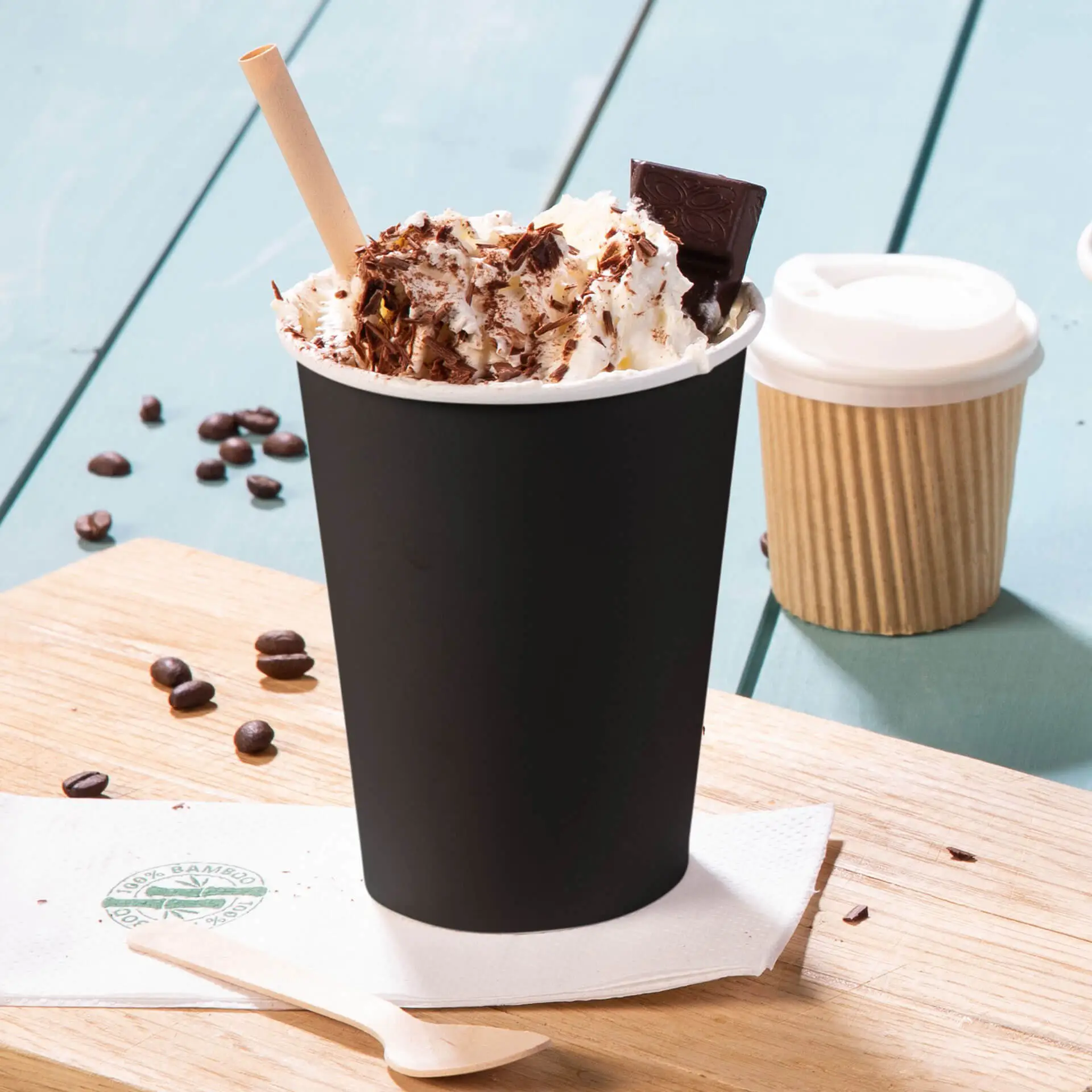 Tazza di carta nera a parete singola con panna montata, scaglie di cioccolato e una cannuccia, servita su un tagliere di legno, con chicchi di caffè nei dintorni.