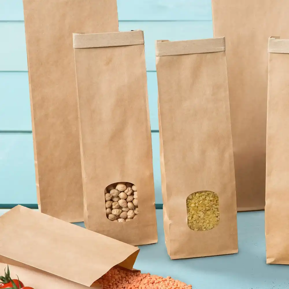 Sacchetti marroni a fondo piatto con finestre trasparenti che mostrano il contenuto, posizionati su una superficie azzurra chiara.