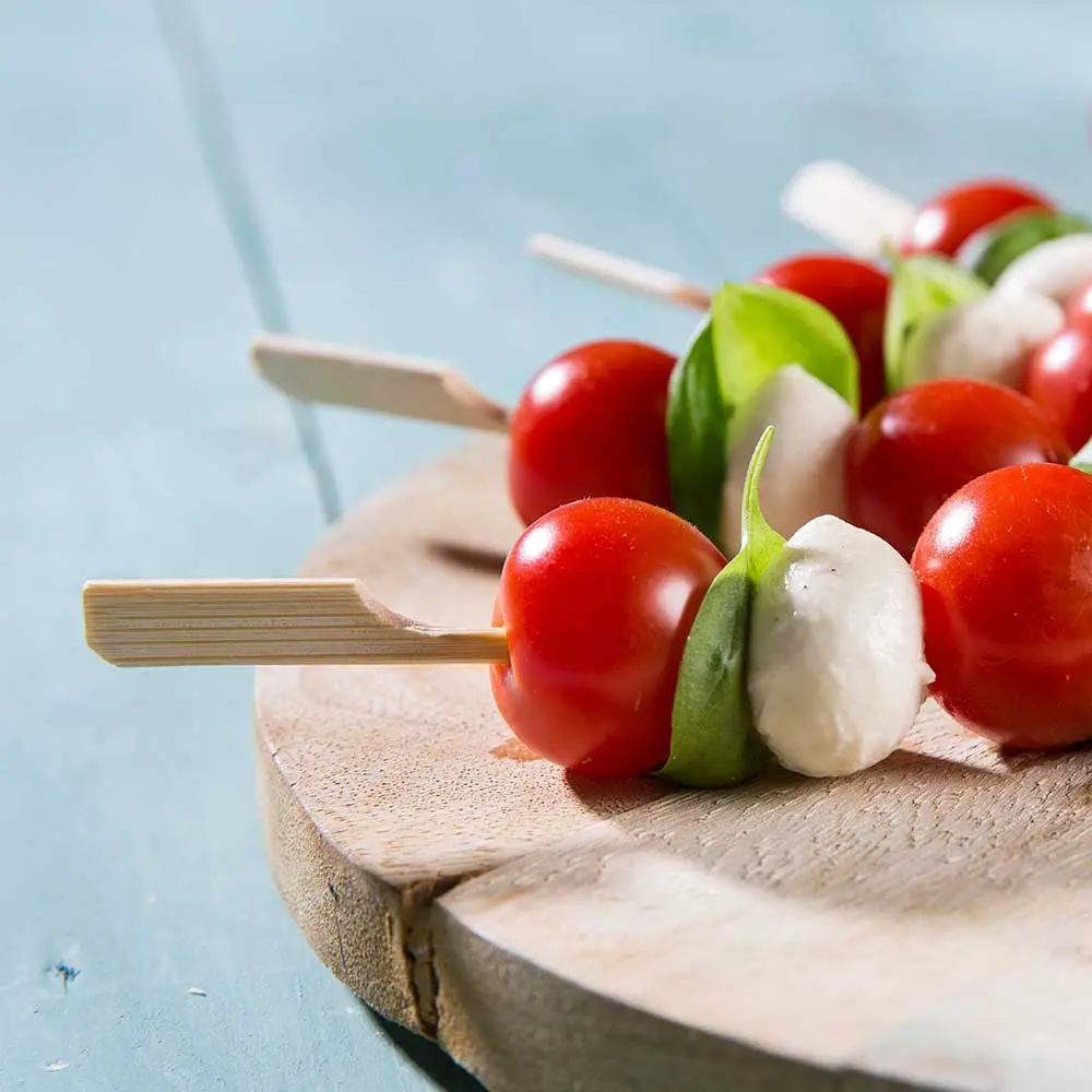 Spiedini con pomodorini, foglie di basilico e palline di mozzarella su un tagliere di legno.