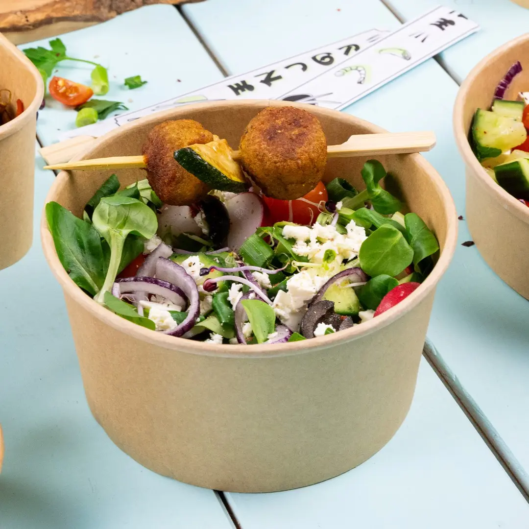 Insalata in una ciotola di carta marrone con verdure, pomodori, cipolle, olive, feta e due falafel su uno spiedino.
