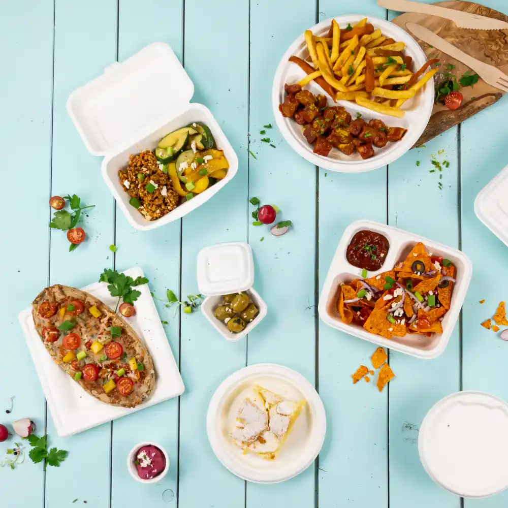 Contenitori alimentari usa e getta assortiti con vari piatti, tra cui patatine, nachos, insalata e dessert su un tavolo di legno blu.