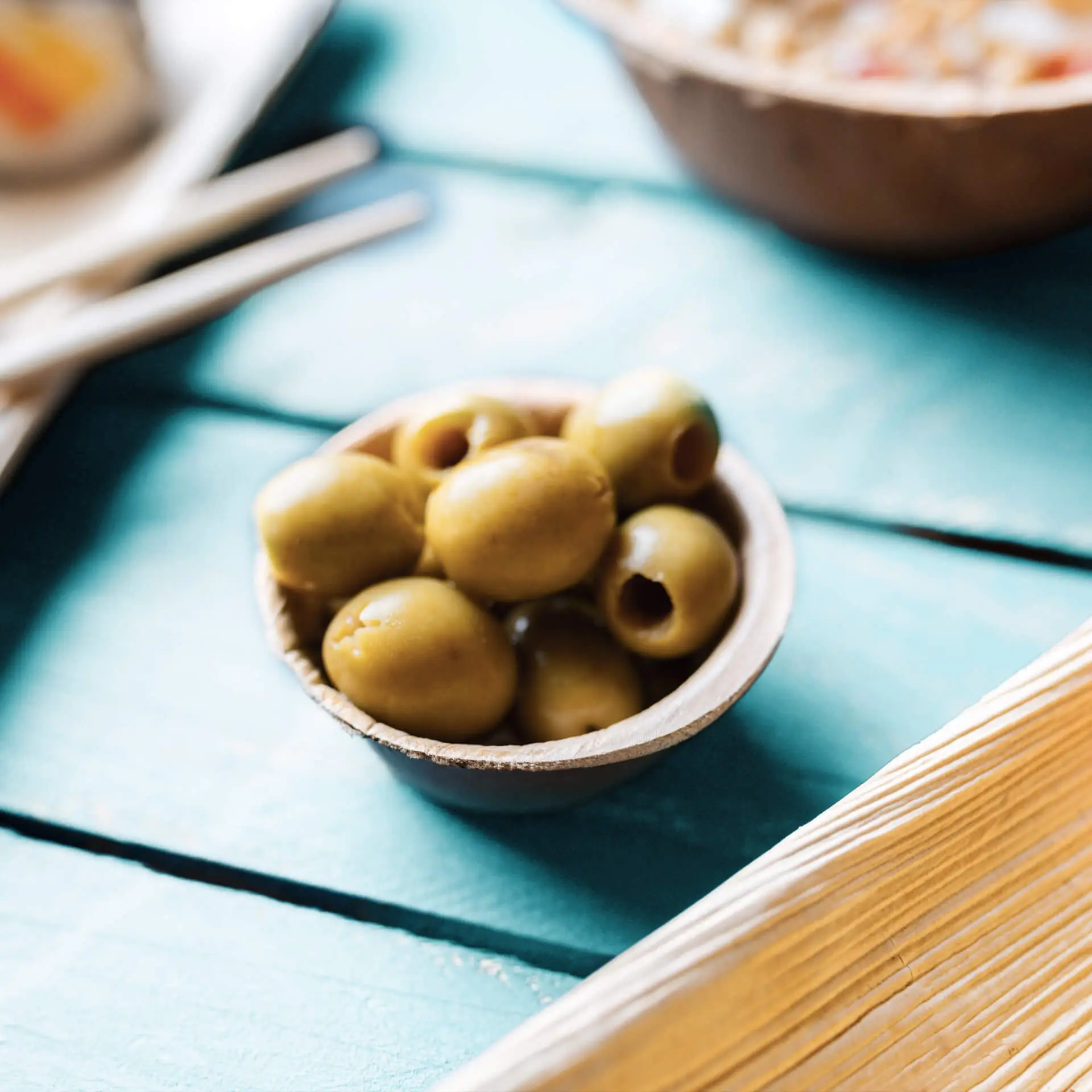 Una piccola ciotola rotonda in foglia di palma piena di olive verdi, posata su una superficie in legno turchese.