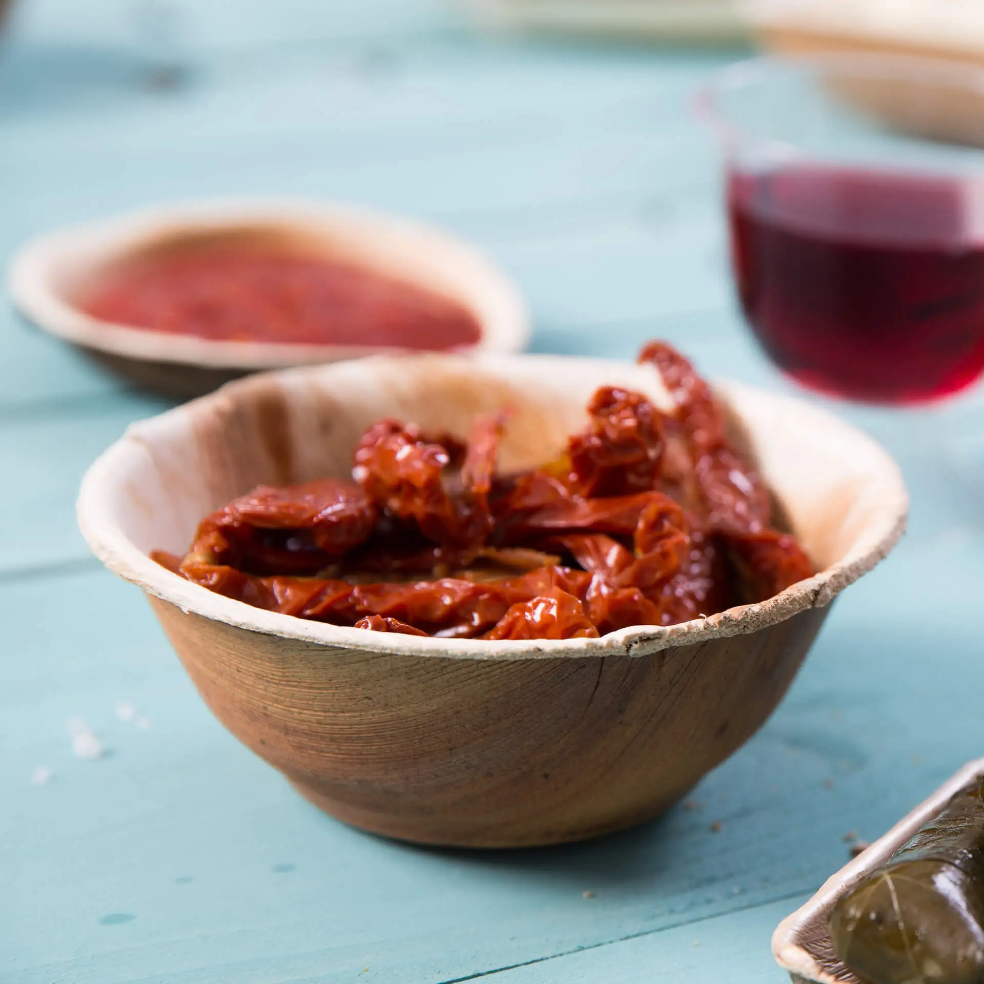 Ciotola di foglie di palma riempita di pomodori secchi al sole; ciotola aggiuntiva e un bicchiere di liquido rosso sullo sfondo.