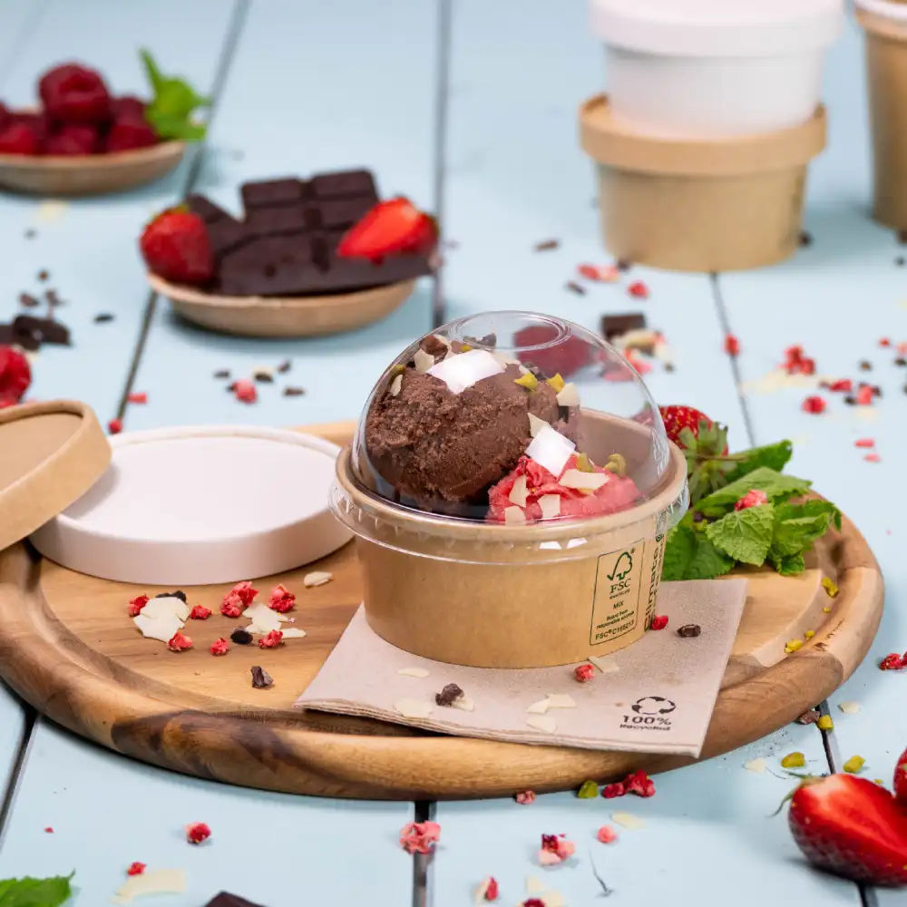 Gelato in un contenitore a cupola trasparente su un vassoio di legno, guarnito con fragole, menta e pezzi di cioccolato.