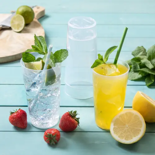 Due bicchieri riutilizzabili con bevande, uno trasparente con una cannuccia contenente lime e menta, e uno giallo con una cannuccia, circondato da fragole fresche e fette di limone.