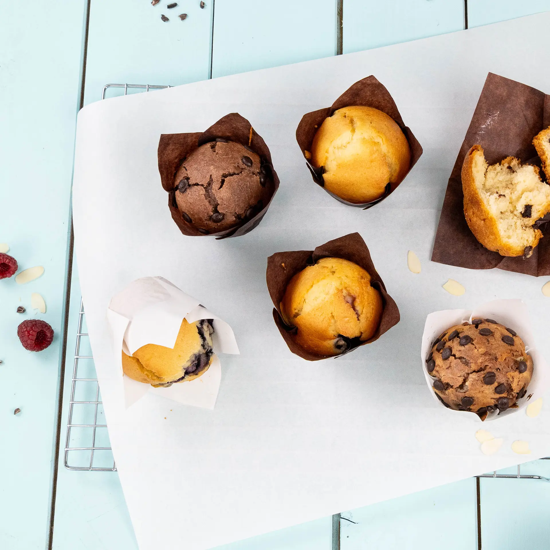Muffin assortiti in bicchieri bianchi biodegradabili su carta da forno; sapori di cioccolato, mirtillo e vaniglia visibili.
