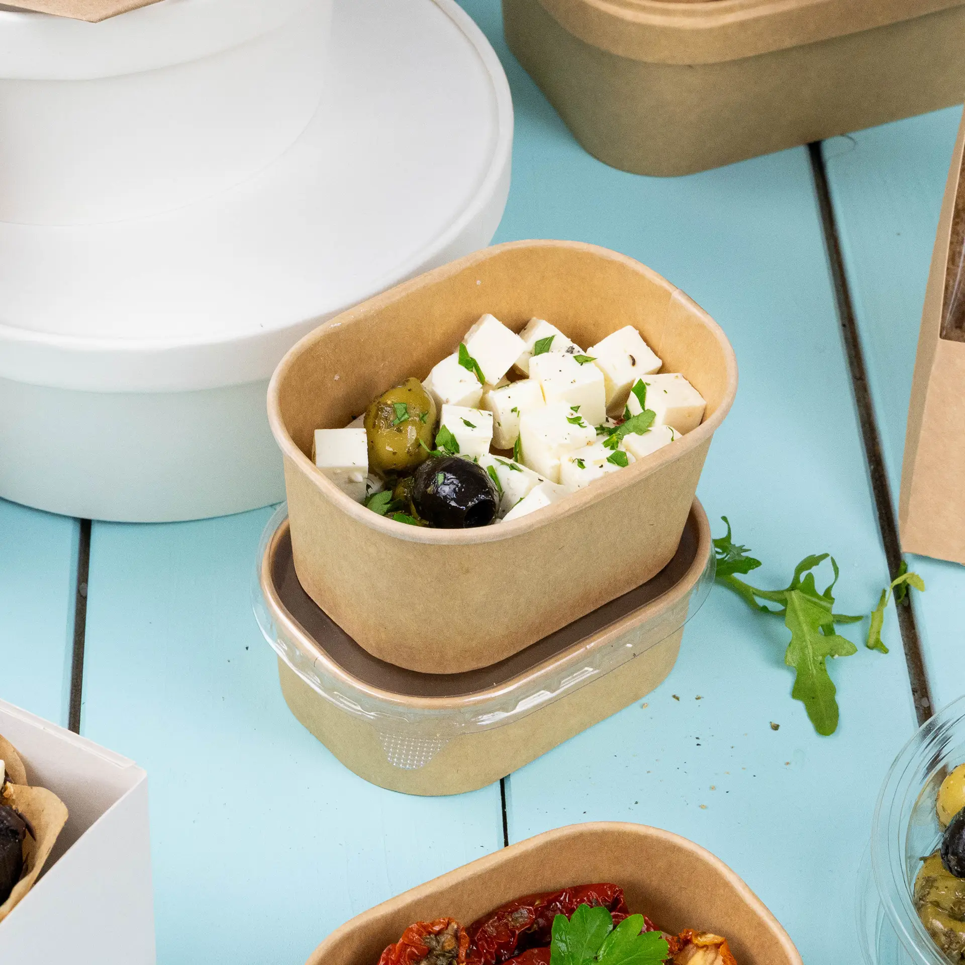 Contenitore alimentare rettangolare in kraft riempito con formaggio a cubetti e olive, con guarnizione di foglie; altri contenitori sono visibili.