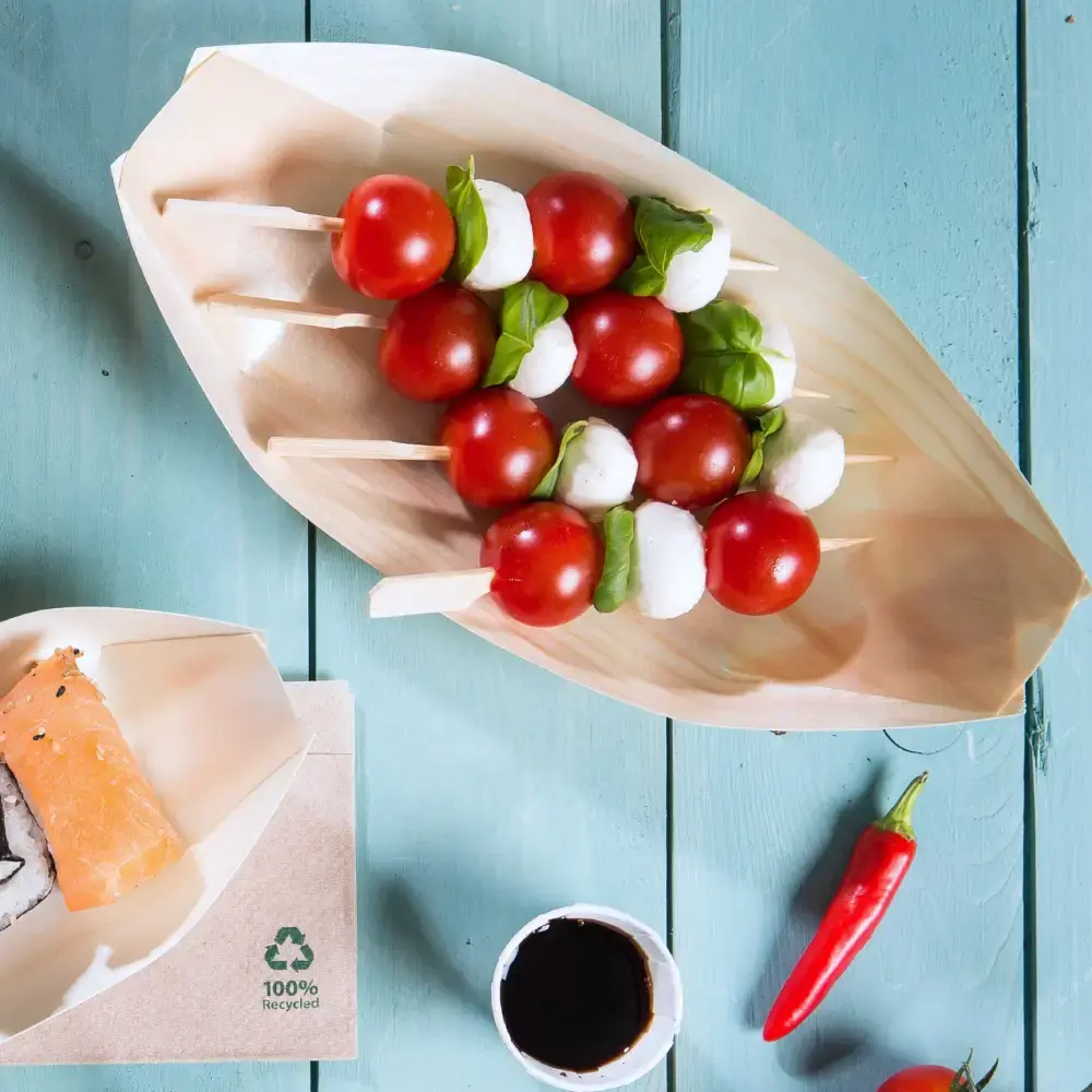 Vassoio in legno a forma di barca con spiedini di pomodorini, palline di mozzarella e foglie di basilico, accanto a sushi e salsa per intingere.
