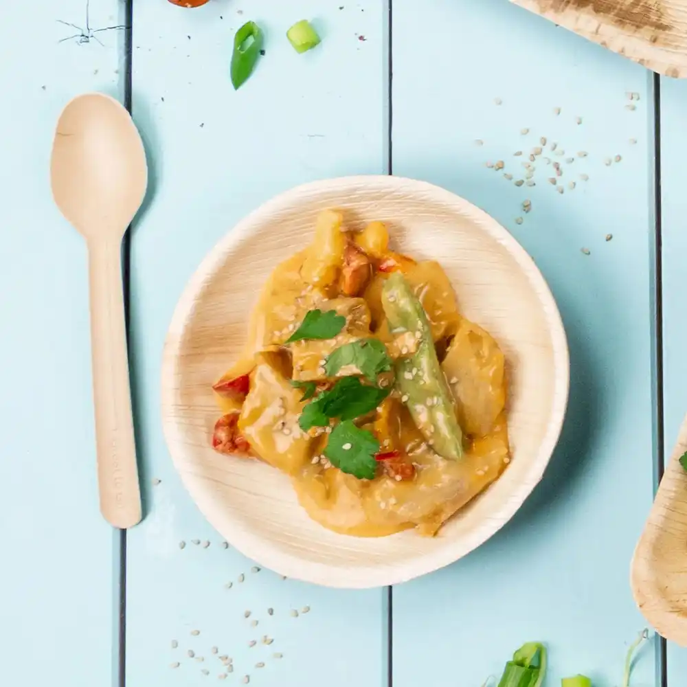 Ciotola in bambù con verdure saltate e salsa, guarnita con semi di sesamo e coriandolo, accanto a un cucchiaio di legno.