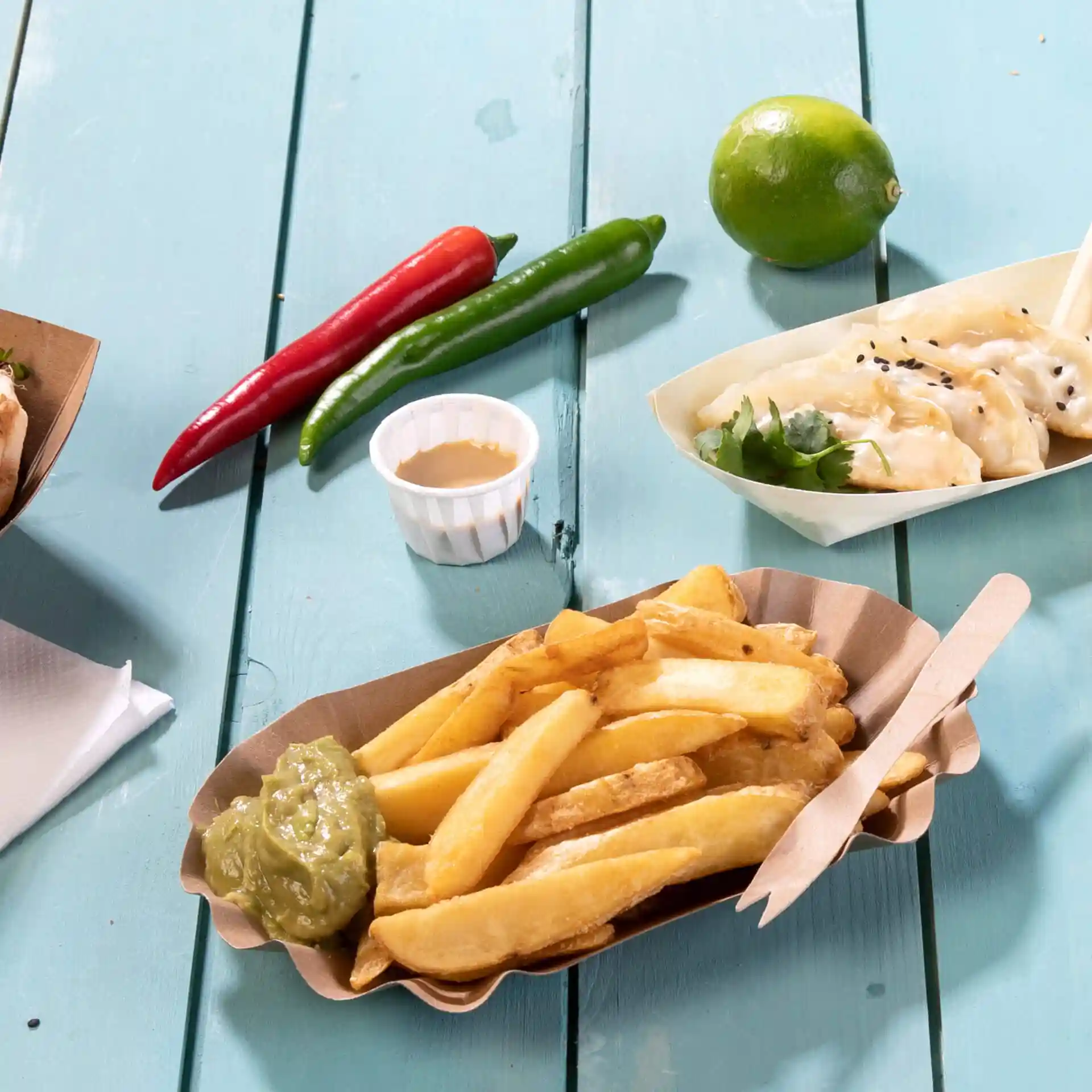 Immagine di una porzione di patatine fritte con salsa verde in un vassoio di carta, circondata da peperoncini, un lime e un piccolo contenitore di salsa.