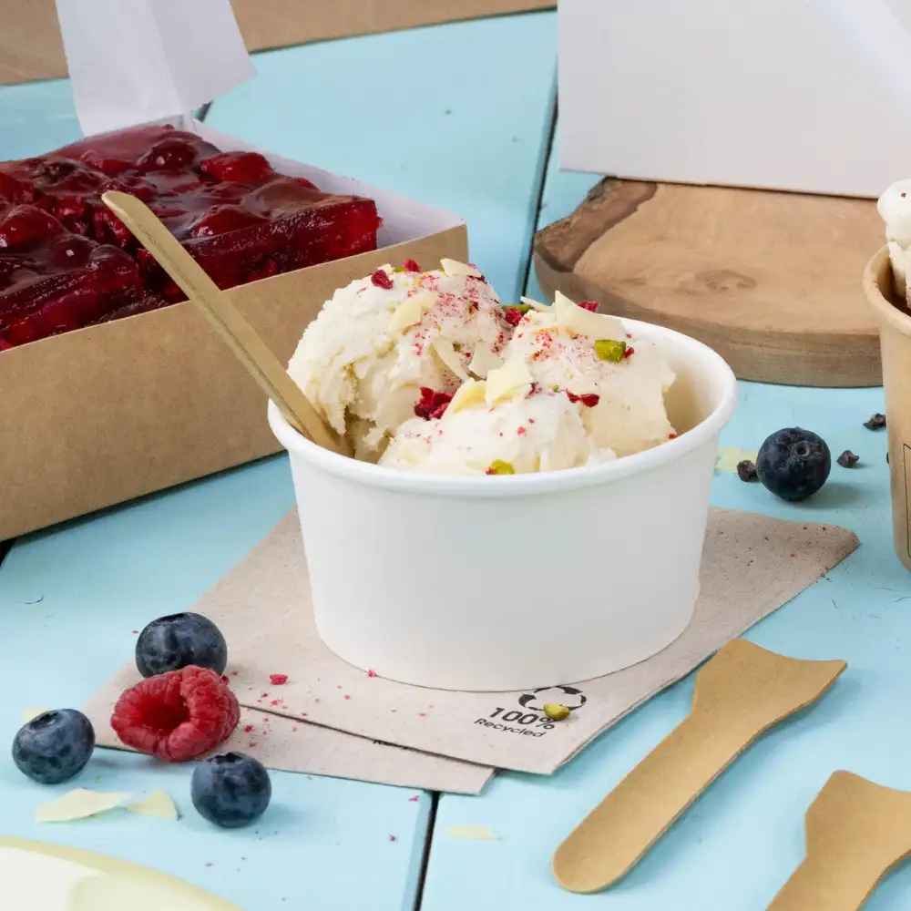 Gelato in un bicchiere bianco, guarnito con noci e frutti di bosco, accanto a una scatola di dessert, con cucchiai di legno e frutta fresca su una superficie blu.