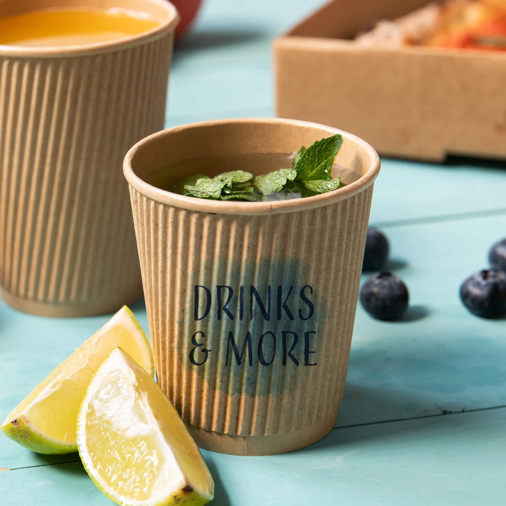 Un bicchiere da caffè da asporto marrone a righe con foglie di menta e spicchi di lime, etichettato "BEVANDE & ALTRO". Altri bicchieri sono parzialmente visibili sullo sfondo.