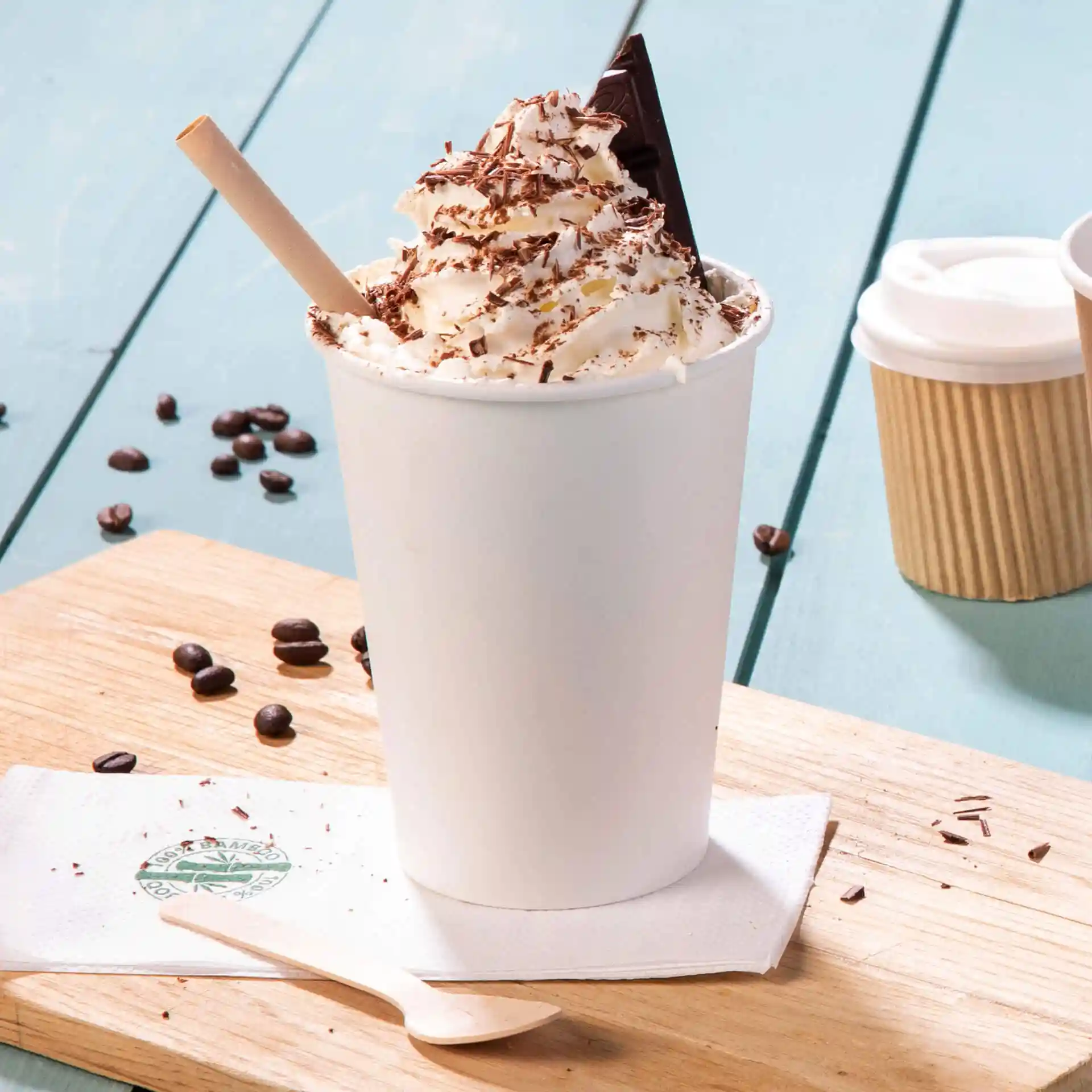 Bicchiere bianco riempito di panna montata, scaglie di cioccolato e un pezzo di cioccolato, con una cannuccia di legno e chicchi di caffè vicini.