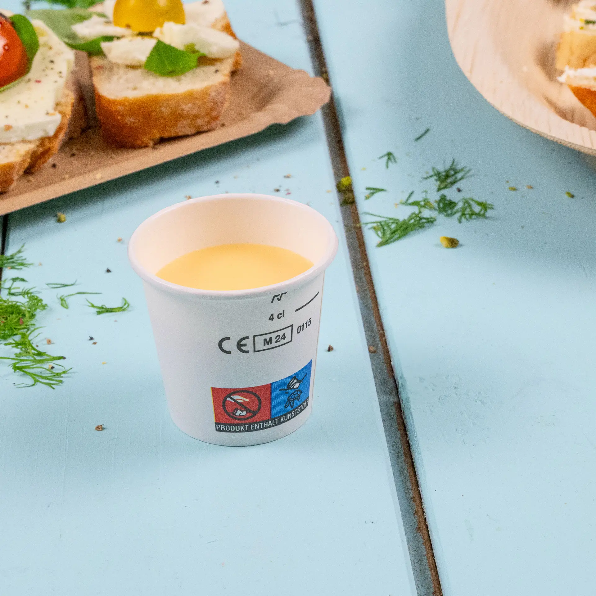 Un bicchiere di carta bianco contenente liquido giallo; accanto ci sono fette di pane con condimenti parzialmente visibili.