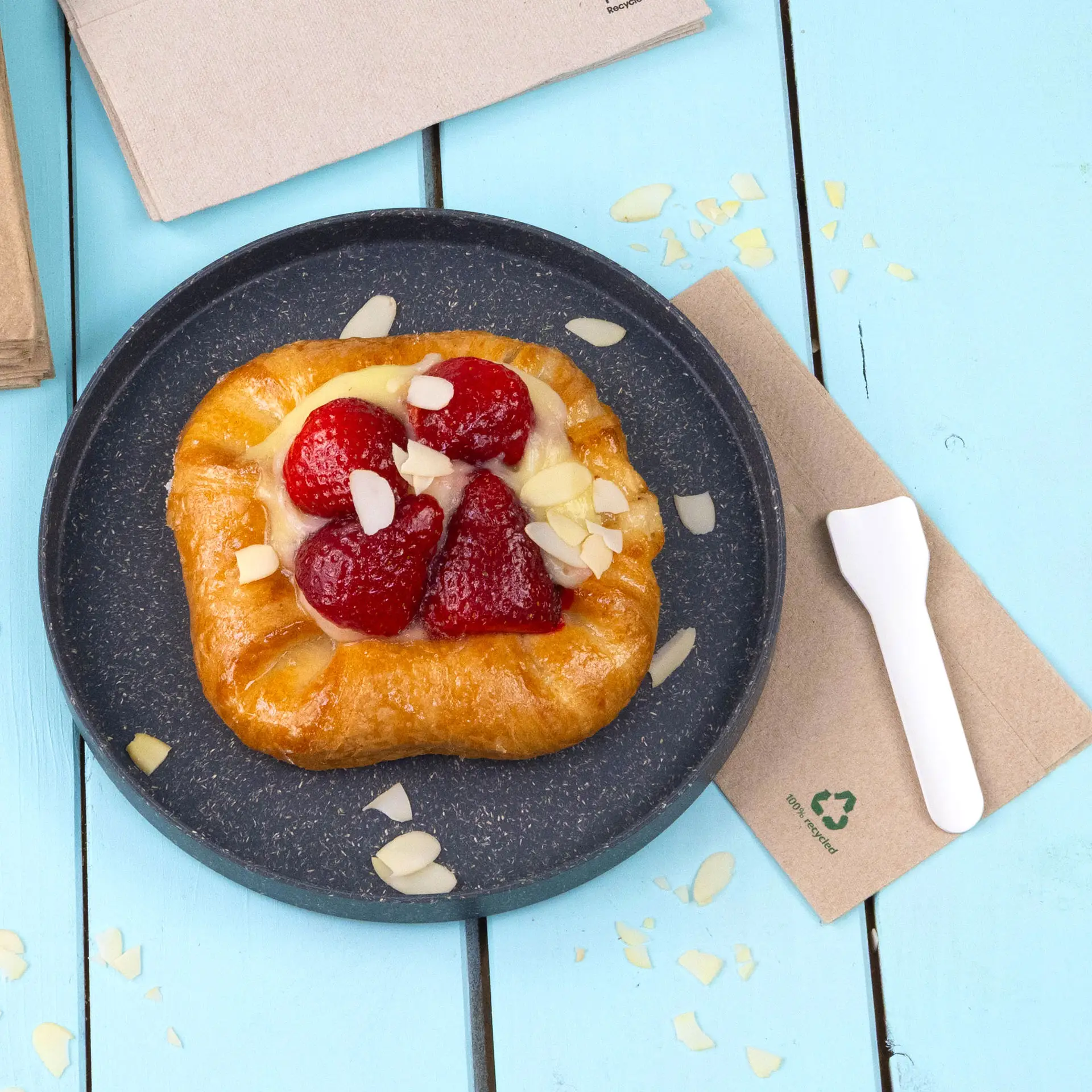 Pasticceria con fragole e fette di mandorle su un piatto scuro, accompagnata da una posata di plastica bianca e un tovagliolo non sbiancato.