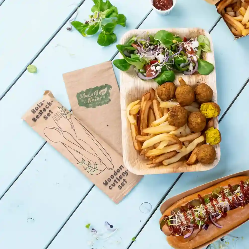 Set di posate in legno accanto a un vassoio di cibo con insalata, patatine, palline di falafel e un hot dog su un tavolo blu.
