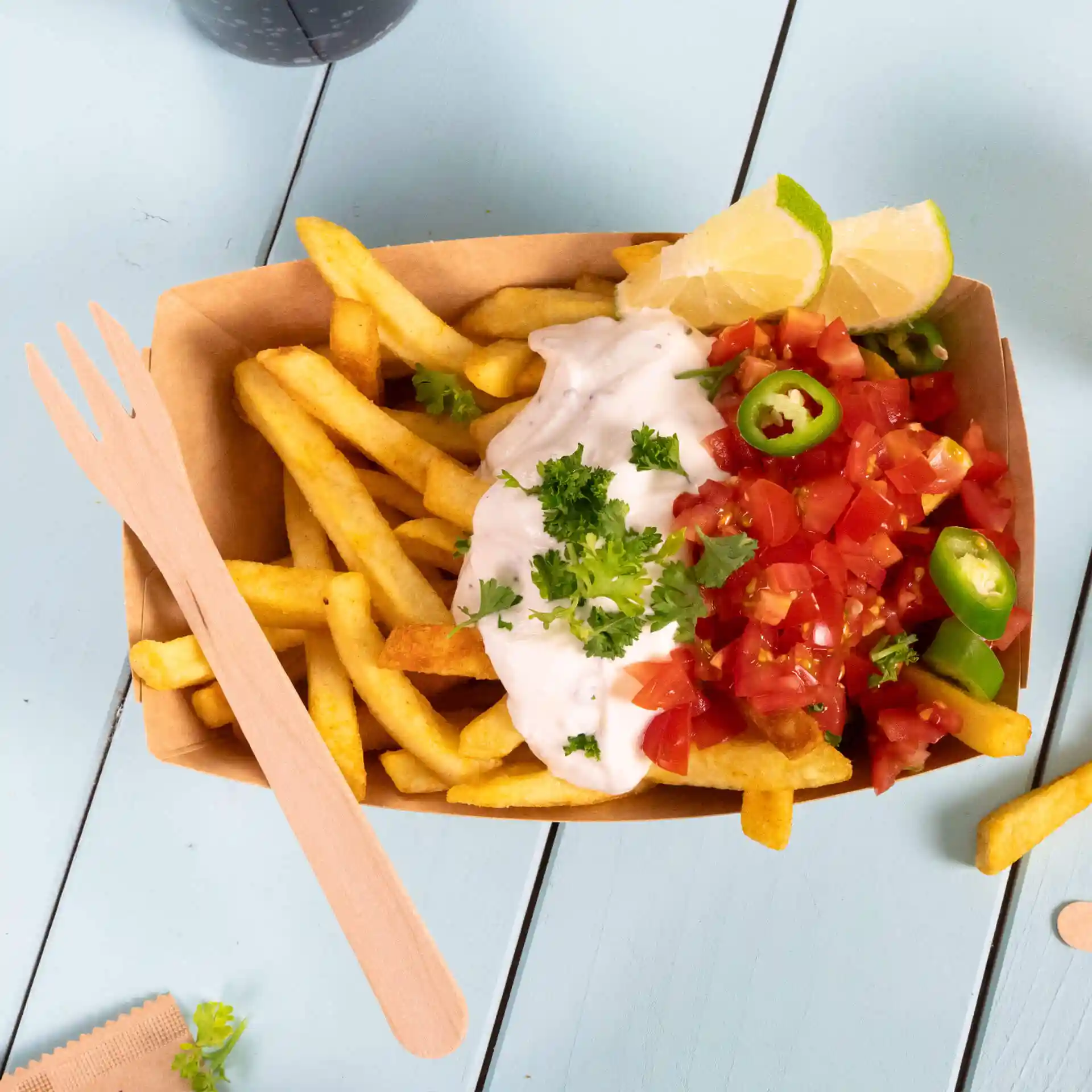 Forchetta di legno accanto a una porzione di patatine fritte guarnite con panna acida, pomodori a cubetti, peperoni verdi e spicchi di lime.