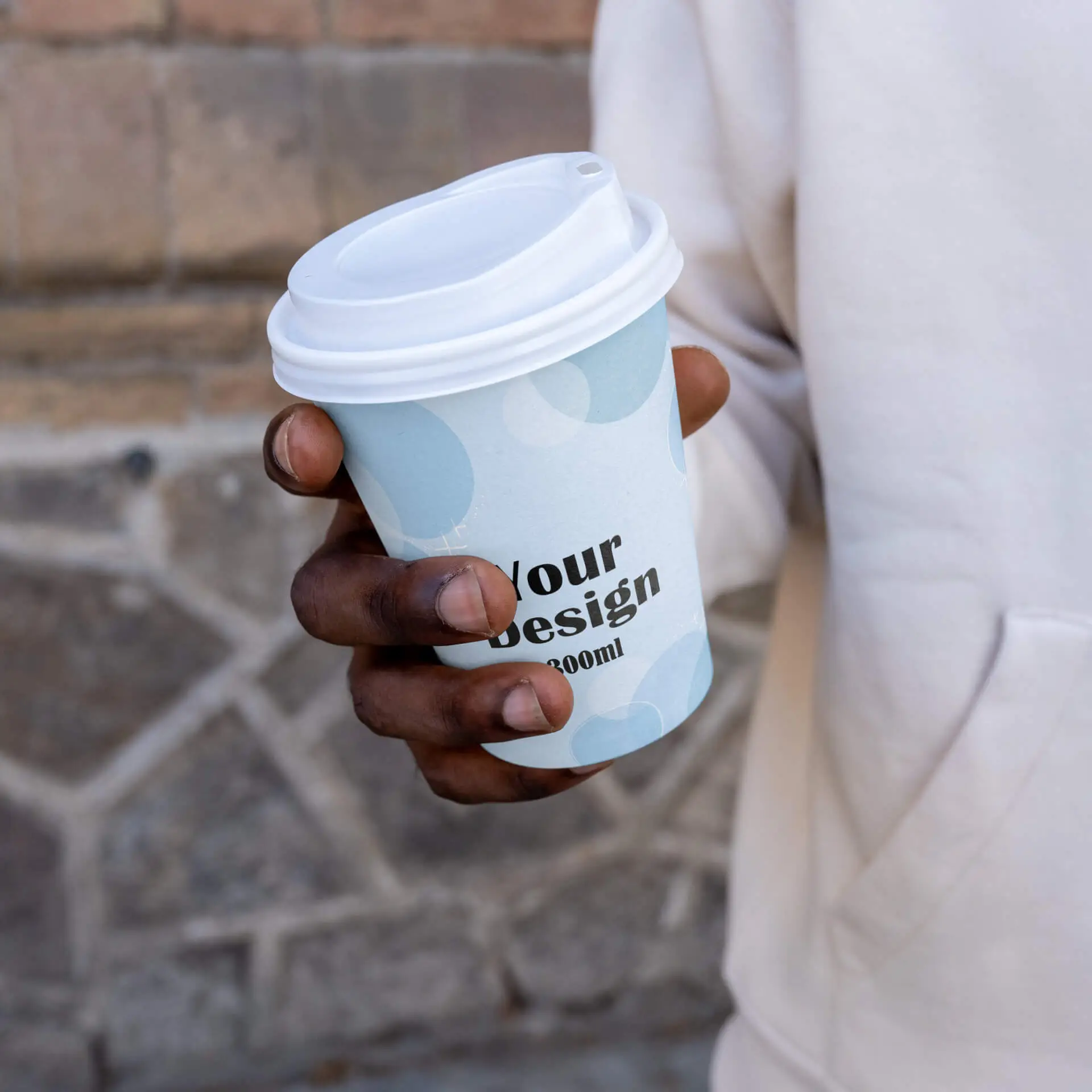 Tazza di carta personalizzabile da 300 ml con design blu e testo nero; tenuta in una mano, su uno sfondo di pietra.