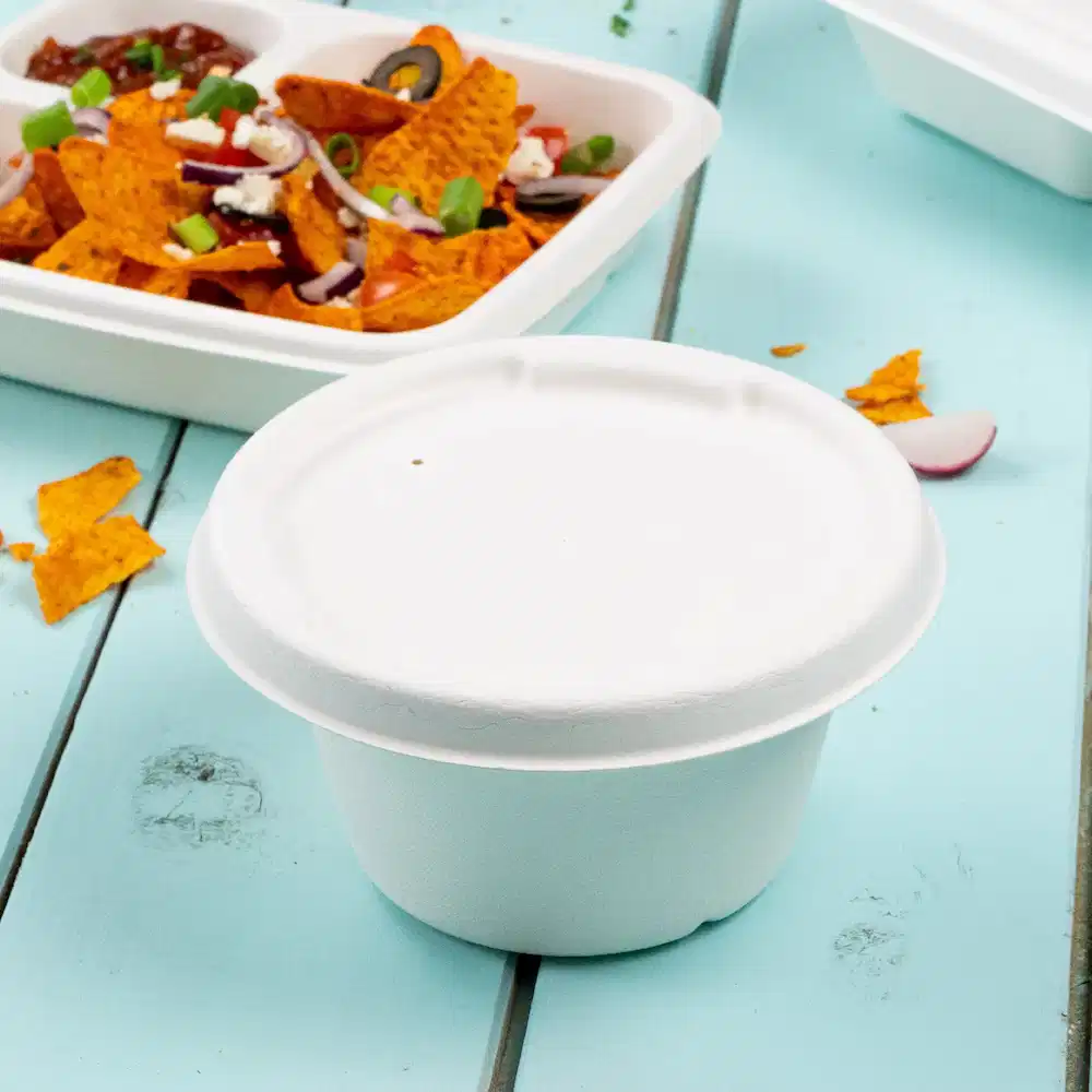 Tazza di carta bianca per zuppa su un tavolo blu con sullo sfondo nachos e un contenitore per alimenti sigillato.