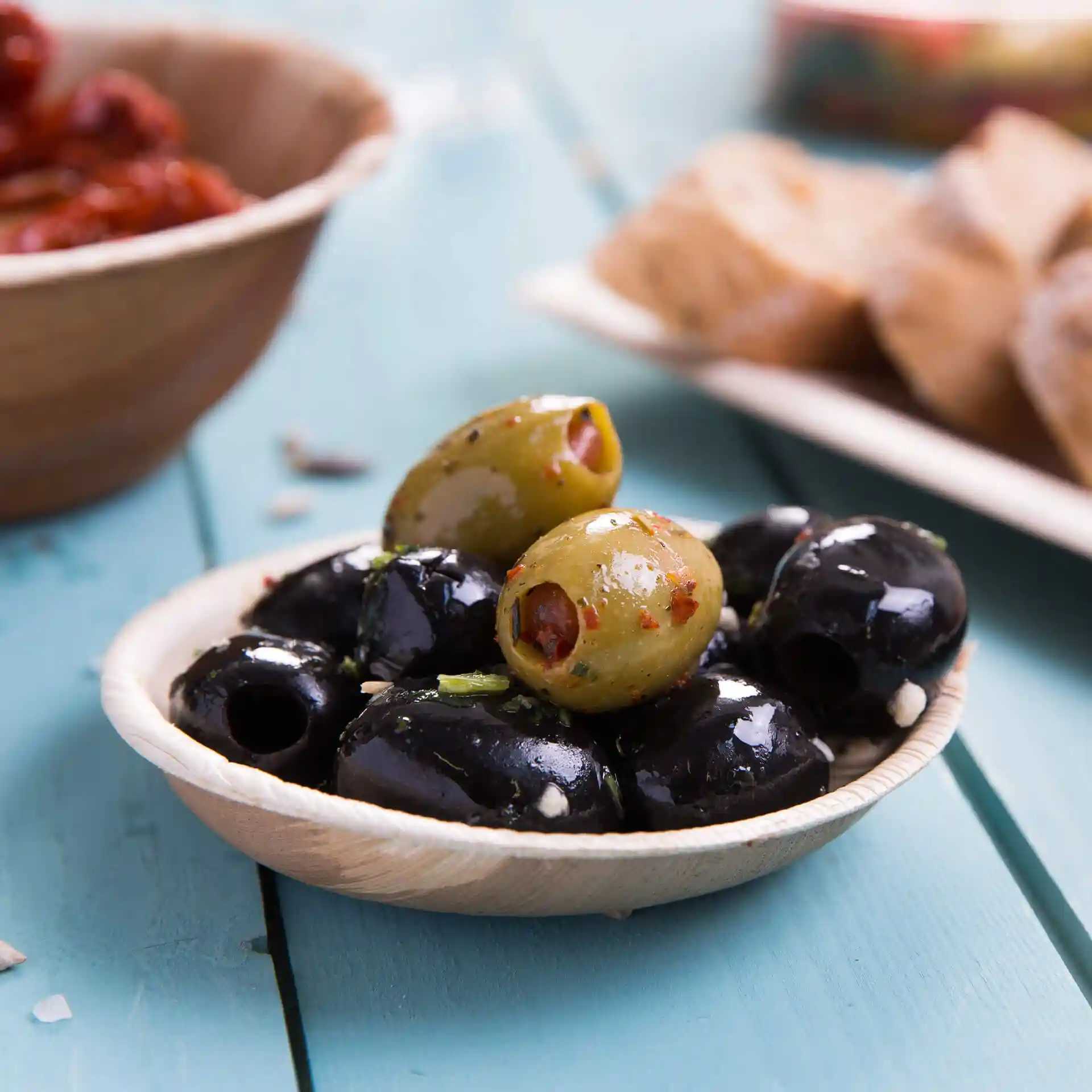 Ciotola in foglia di palma contenente olive nere e verdi, posata su una superficie di legno blu.